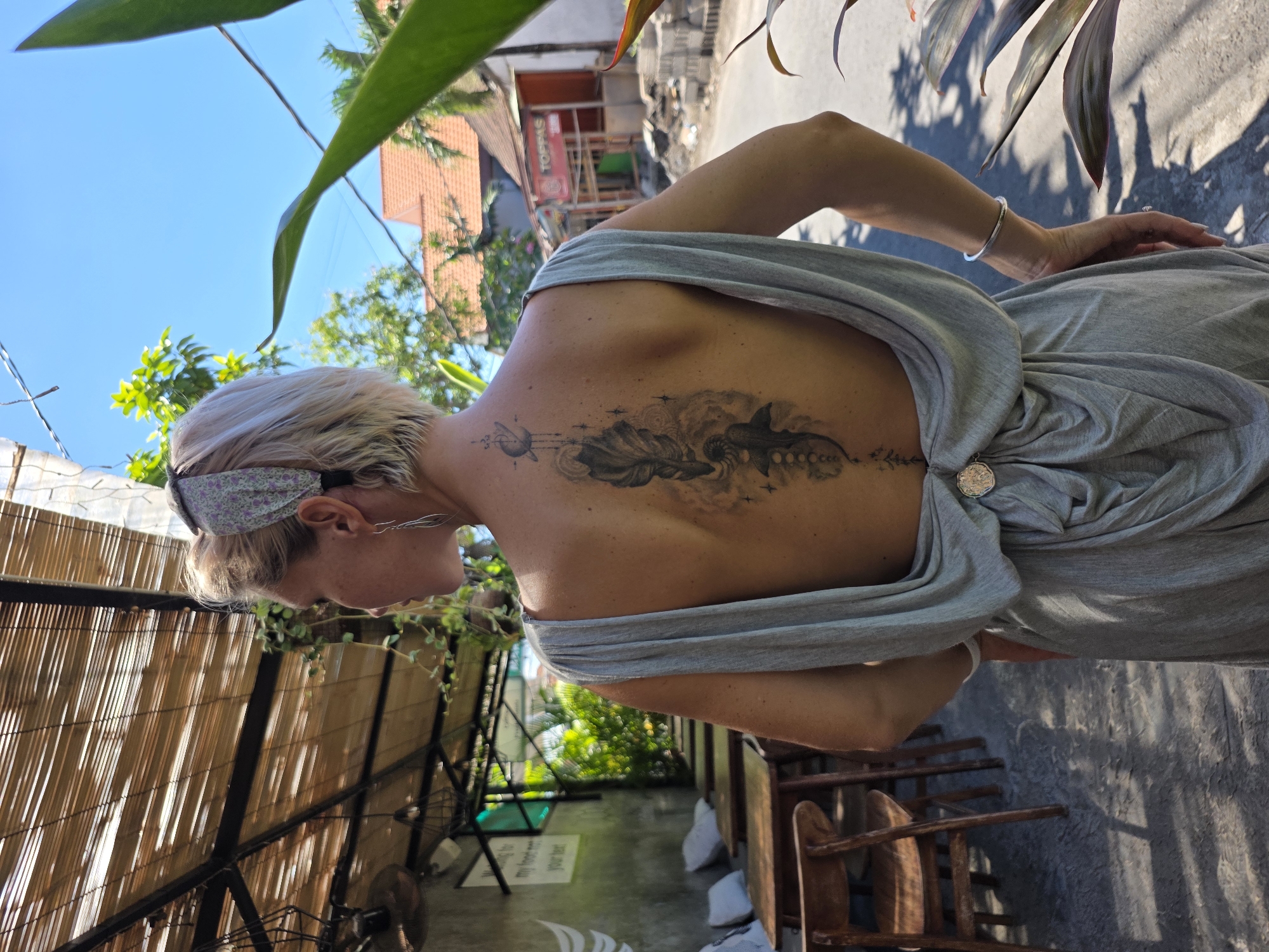 A medium shot depicts the upper back of a woman with short, blonde hair and a patterned headband, as she stands outside in what appears to be North Kuta, Indonesia. She wears a light gray dress that drapes around her shoulders and upper back, secured at the lower back with a decorative broach or clasp. A prominent tattoo extends along her spine. The tattoo design includes celestial and symbolic imagery. The background shows an outdoor scene with greenery, buildings including a structure with a "TOPPING" sign, and a shadowed street area. To the left, a bamboo structure and additional foliage are visible, adding to the tropical outdoor setting.