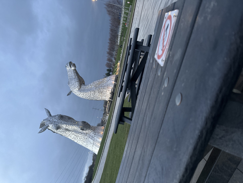 The image displays an outdoor scene featuring The Kelpies, two colossal metallic horse head sculptures, under an overcast, dark grey sky. The location is The Helix Park, Falkirk, Scotland, United Kingdom.
In the immediate foreground, a dark-colored wooden picnic table occupies the lower portion of the frame, angled upwards from left to right. Visible on its slatted surface are several small, round, silver-colored metallic objects, and a white square sticker with a red circular border enclosing a diagonal line through a lit cigarette, indicating a "No Smoking" prohibition.
Behind the picnic table, a paved walkway leads towards the bases of the two horse sculptures. The sculpture on the left, depicted with its head slightly raised and facing left, is partially obscured by the table's perspective. The sculpture on the right presents a horse head looking straight ahead with its mouth slightly open. Both sculptures are composed of numerous polished metal plates and appear illuminated from below.
The midground features green grassy areas and paved pathways surrounding the sculptures. In the background to the left, a grassy slope rises, with power lines and a large metal pylon visible against distant trees. To the right, a row of slender, light-barked trees extends horizontally, bordering what appears to be a canal or water feature, with additional distant structures and a street light also visible. No people are present or identifiable in the scene. There are no explicit actions or interactions depicted.