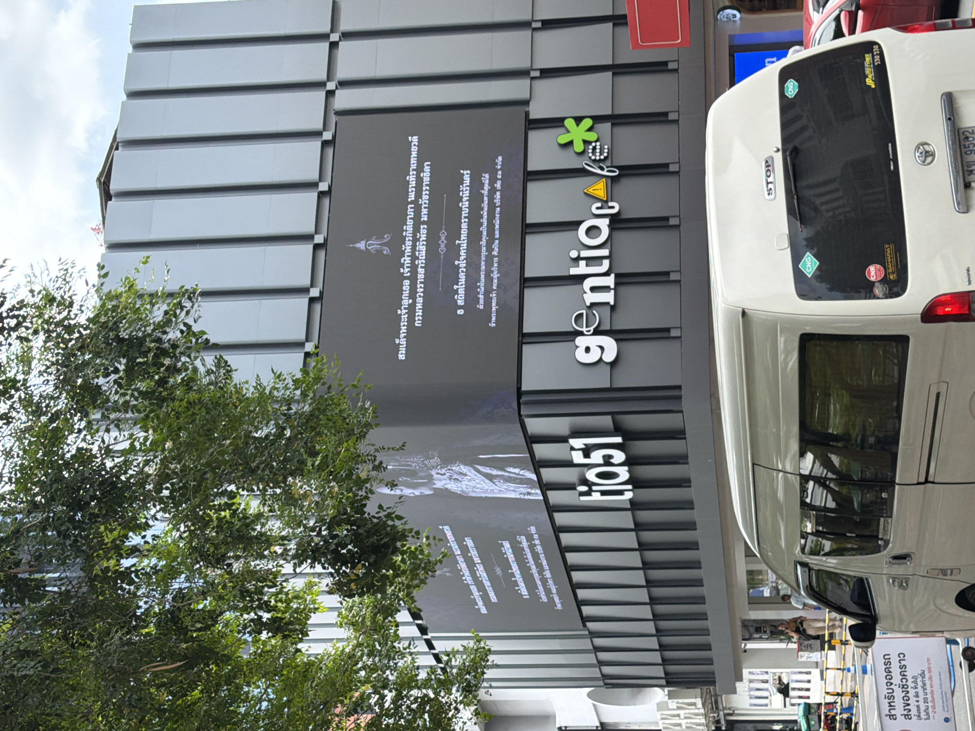 The image shows a street scene in Bangkok, Thailand, in front of a modern building with a large digital display. The building is predominantly light gray and features vertical paneling. On the lower part of the building, there is signage that reads "tia51" and "gentia cafe" with a green asterisk logo.
The digital display on the upper part of the building shows an image of a woman in traditional attire and Thai text. Below this, there is additional Thai text.
In the foreground, a white van is parked on the street. The van has several stickers on its rear window, including "CNG," "CNR," "SAHARAT," and "JP AUTO TYRES." There is also a license plate visible on the van.
Further down the street, a red car is partially visible. Other elements of the urban environment include trees with green foliage, street signs, and poles with yellow and white barriers. Two individuals are walking on the sidewalk in the background. The sky is partly cloudy. The overall scene depicts a typical day in a commercial district of Bangkok.