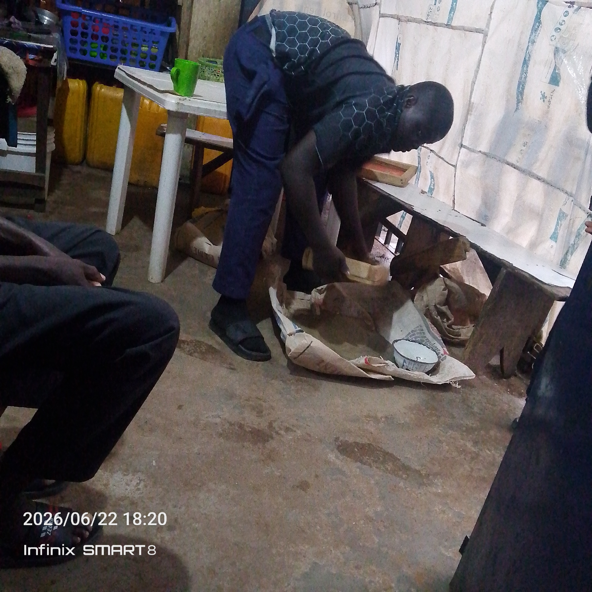 The image shows a group of people in what appears to be a storage or workshop area. In the foreground, a person is sitting on the floor, wearing dark pants and black sandals. To their left, another person, wearing a black t-shirt with a hexagonal pattern and dark blue pants, is bending over a table and interacting with some objects. The table is white with white legs and is placed on a concrete floor. Behind the table, there are yellow plastic containers and a blue basket. In the background, there are large rolls of what looks like white material, possibly canvas or insulation, with blue markings. A small bowl and some crumpled paper or fabric are on the floor near the bending person. The lighting suggests it might be indoors or late in the day.
Visible text in the image includes a timestamp "2026/06/22 18:20" and "Infinix SMART 8" at the bottom left corner. There are also some faint markings on the white material in the background, which are difficult to decipher. The overall scene is somewhat cluttered and utilitarian, suggesting a workspace or storage facility. No specific emotions are discernible from the subjects.