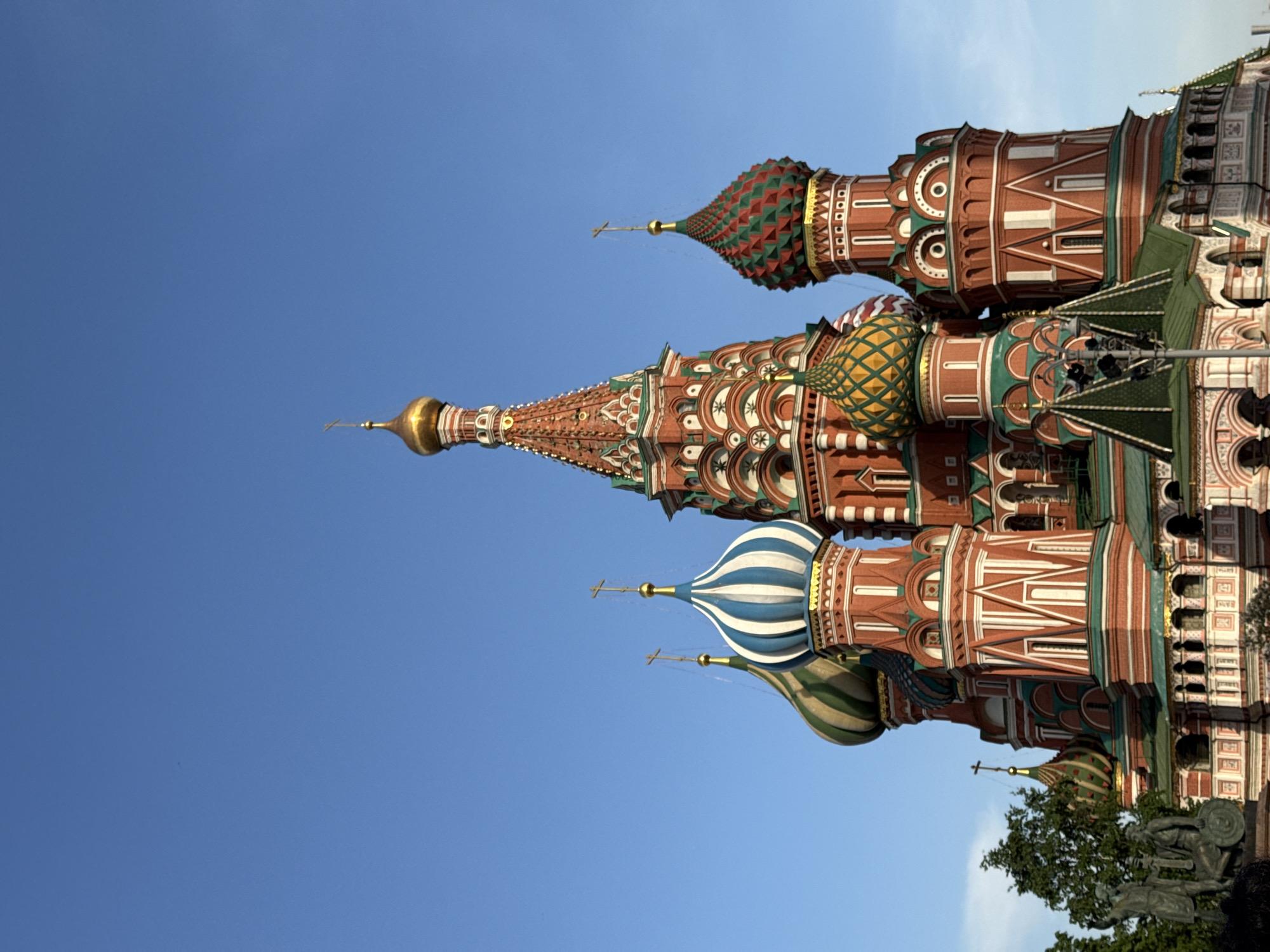 The image features St. Basil's Cathedral in Moscow, Russia. The cathedral's distinctive onion-shaped domes are prominent, showcasing colorful patterns in red, green, gold, and blue. The sky is clear and blue. The structure is composed of brick with intricate designs. At the bottom right of the image, a portion of a monument and some trees are visible.