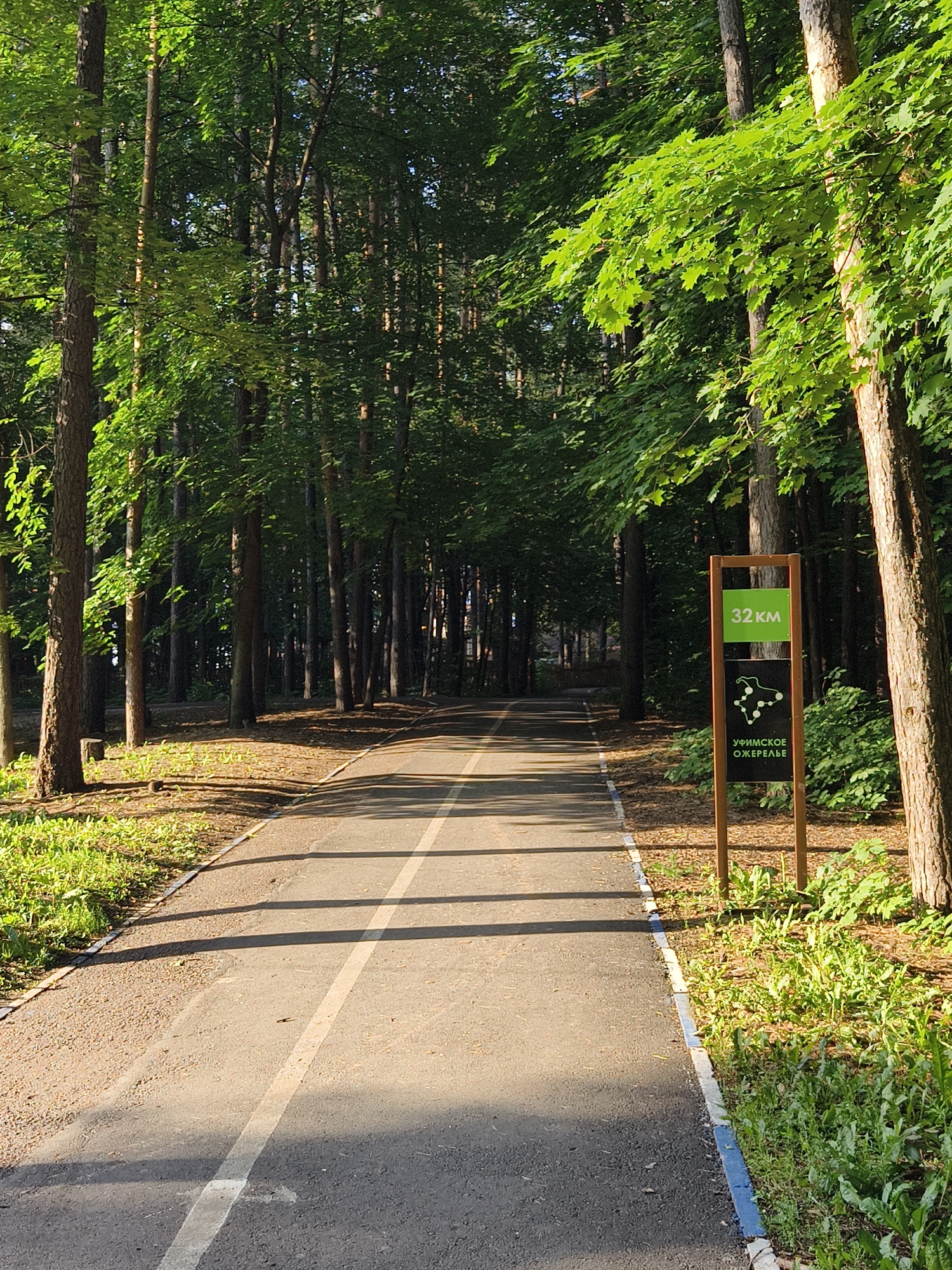 A paved path, likely a bicycle or running trail, winds through a sun-dappled forest. Tall trees with lush green foliage line both sides of the path, casting long shadows. A wooden signpost stands on the right side of the path, featuring a green rectangular panel with "32 KM" printed in white text. Below this, a black rectangular panel displays a white graphic of a stylized animal or object with connected dots, and the text "УФИМСКОЕ ОКЕРЕЛЬЕ" (Ufimskoe Oskerele) in white Cyrillic letters, indicating this location is part of the Ufa Belt Nature Reserve in Ufa, Russia. The path has a white line painted down its center. The ground beside the path is covered in grass, small plants, and fallen leaves, with some wood chips visible around the base of the signpost. The scene appears to be set on a clear, sunny day. There are no people or animals visible in the frame.