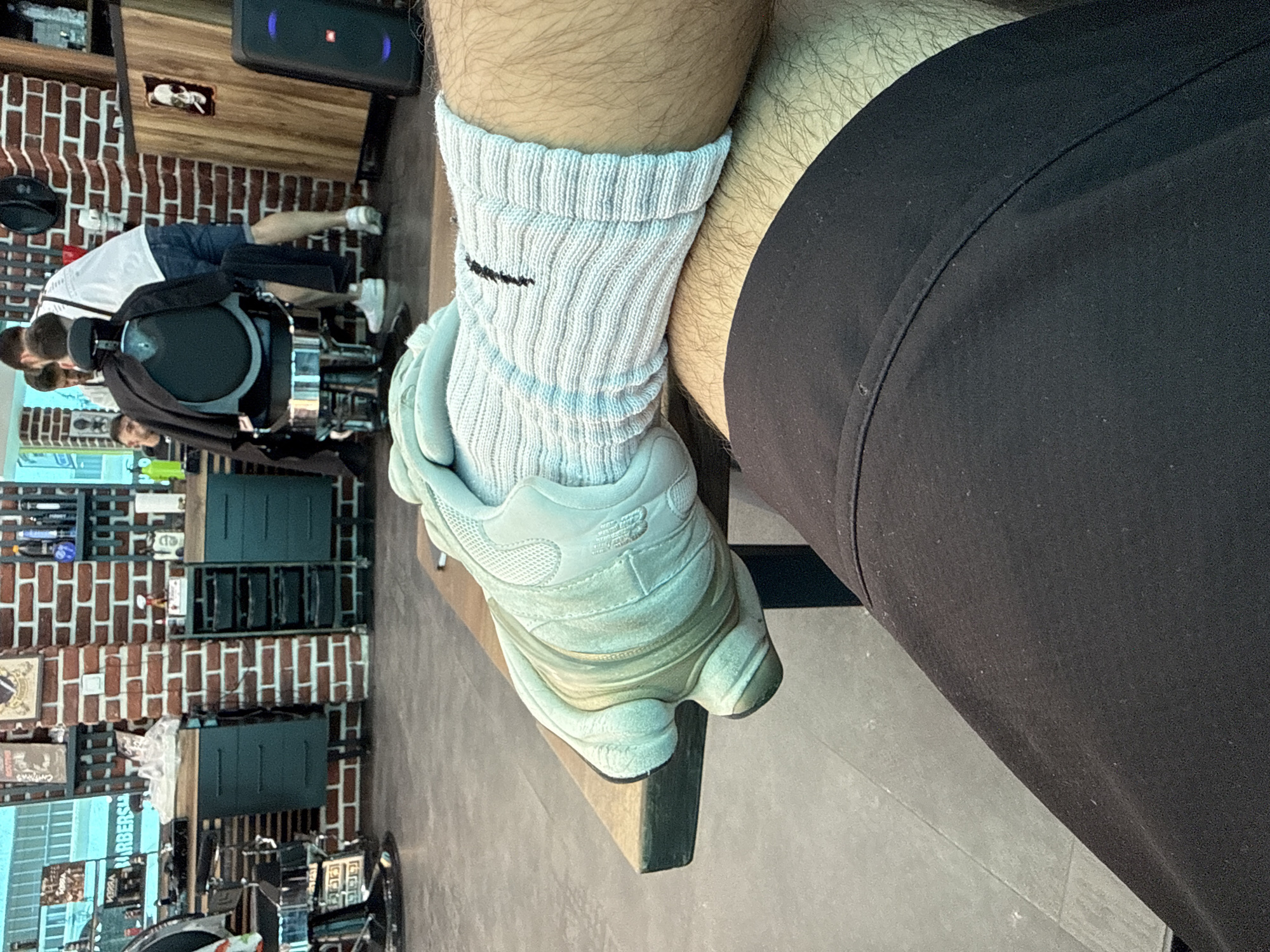 The image depicts an interior scene, likely a barbershop, with brick walls and various styling stations. In the foreground, a person's leg, covered by black shorts and a white sock, is resting on a wooden table. A light-colored athletic shoe is on the person's foot. In the background, several individuals are present, with one person seated in a barber chair, appearing to be receiving a haircut from another person. The overall atmosphere suggests a busy, functional establishment. This scene is located in Shumen, Bulgaria.