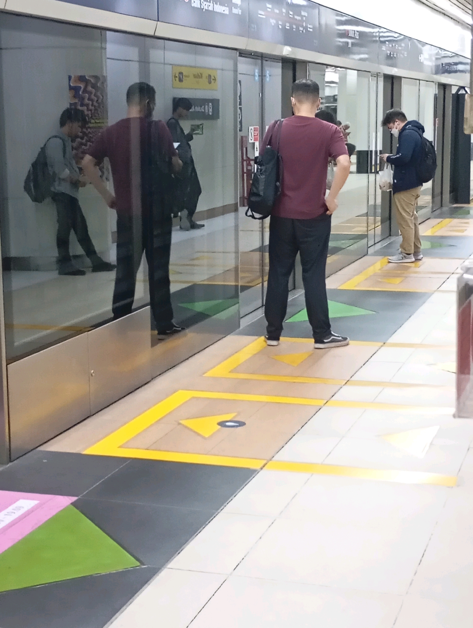 The image shows a platform at a train station in Central Jakarta, Indonesia. Three individuals are visible. In the foreground, a person wearing a maroon t-shirt and black pants stands facing away from the camera, looking towards a train door. To their left, another person in a grey shirt and black pants leans against a wall, also facing away from the camera. To the right of the person in the maroon shirt, a third person wearing a dark blue jacket and khaki pants stands looking at their phone. The platform features yellow and grey floor markings, including directional arrows indicating a queuing system. A reflective glass barrier separates the platform from the train tracks.