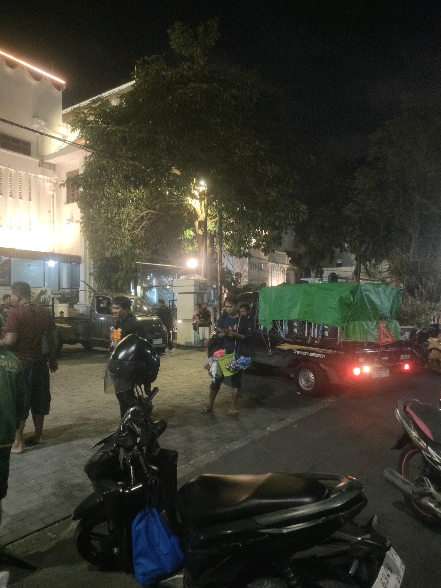 The image depicts a nighttime street scene in Surabaya, Indonesia, likely an urban area with commercial activity. In the foreground, two dark-colored scooters are parked, one with a blue bag hanging from its handlebars. The ground is paved with cobblestones.
In the mid-ground, several people are visible. A man in a red shirt and black shorts stands to the left, looking away from the camera. To his right, a man in a black shirt is seen from the side, smiling. Further right, a pickup truck is parked, its cargo covered with a green tarp. The truck has its taillights illuminated. Another person, dressed in a blue shirt, is standing next to the truck, holding a basket of what appears to be flowers or candy. Several other individuals are seen in the background, some standing near a building.
The environment includes a building on the left side of the image, illuminated by artificial lights. Trees are present throughout the scene, their foliage dark against the night sky. Streetlights and building lights create a moderately lit atmosphere. The overall impression is of a busy street or marketplace at night.