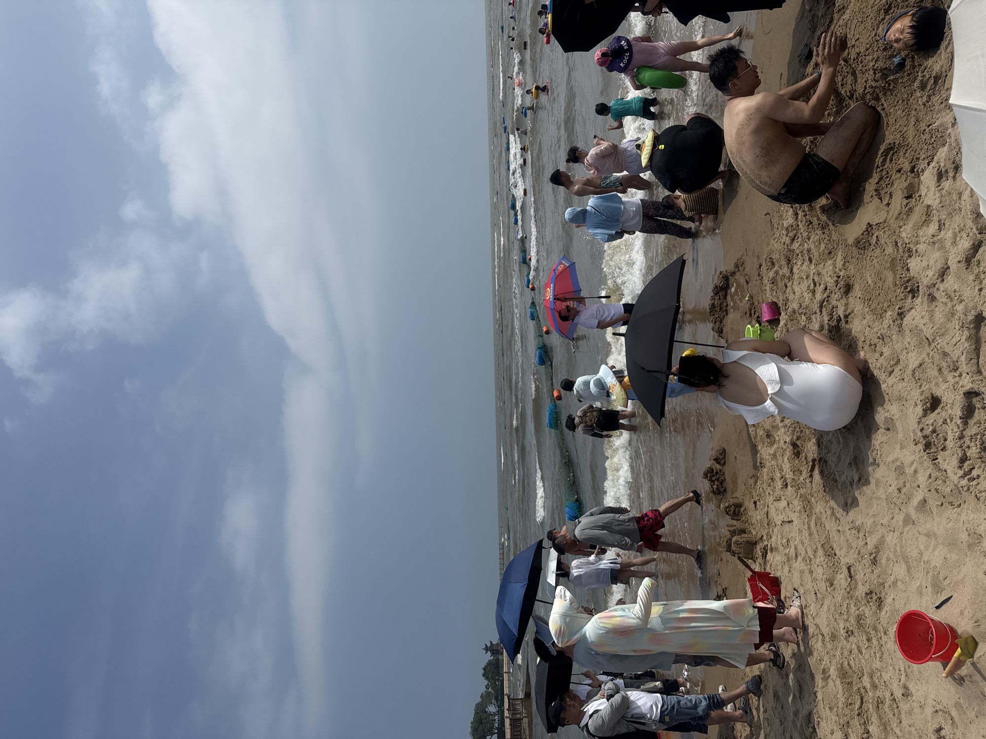 The image depicts a bustling beach scene in Qinhuangdao, China, likely during a sunny day with some cloud cover, indicated by the presence of many people using umbrellas. The tide appears to be coming in, with moderate waves breaking on the shore. The sand is a light brown color, and in the foreground, there are several sandcastles being built, along with children's toys like a red bucket and a yellow shovel.
Numerous individuals are present, dressed in swimwear and casual beach attire. Some are wading in the shallow water, while others are standing on the wet sand or further up on the dry sand. A diverse range of activities is observed: people are holding umbrellas for shade, playing in the sand, and socializing. In the water, there are visible buoys or floats, suggesting a designated swimming area.
In the mid-ground, several people are standing in the water, some holding umbrellas. Further out in the sea, more people are visible, swimming or enjoying the water. The background shows a hazy horizon with the sea extending to meet the sky. In the far distance, a structure resembling a pavilion or temple can be seen on the left side of the frame, hinting at the coastal landscape surrounding the beach. The overall impression is one of a popular public beach on a warm day.