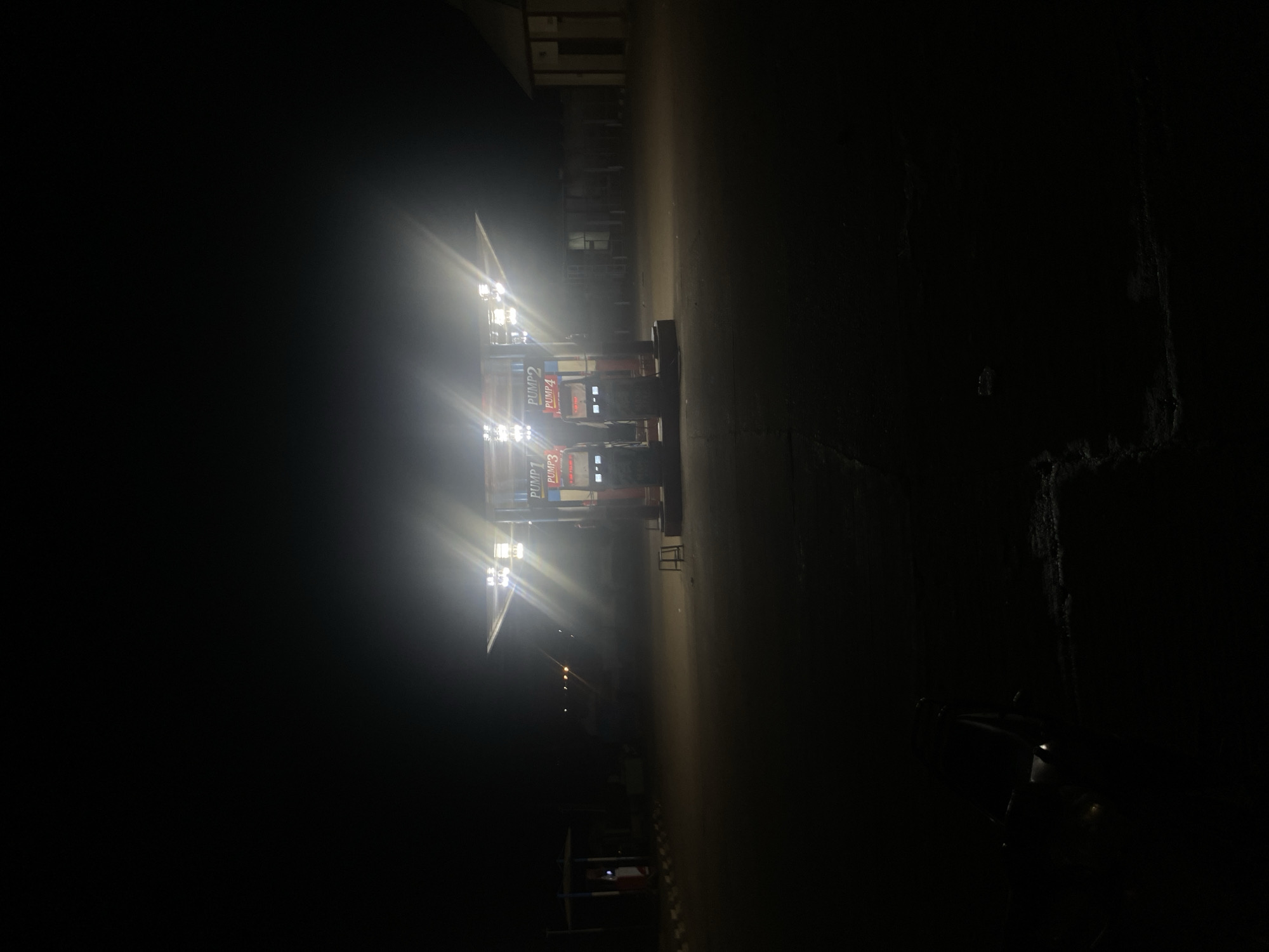 At night in Tukur Tukur, Nigeria, a brightly lit gas station stands out against the darkness. The pumps are illuminated, with "PUMP 1," "PUMP 2," "PUMP 3," and "PUMP 4" marked on them. The area immediately around the station is lit, contrasting sharply with the darker surroundings. A motorcycle is parked on the right side of the image, partially obscured by the darkness. Other structures are visible in the background, but details are hard to distinguish. The ground appears to be paved.