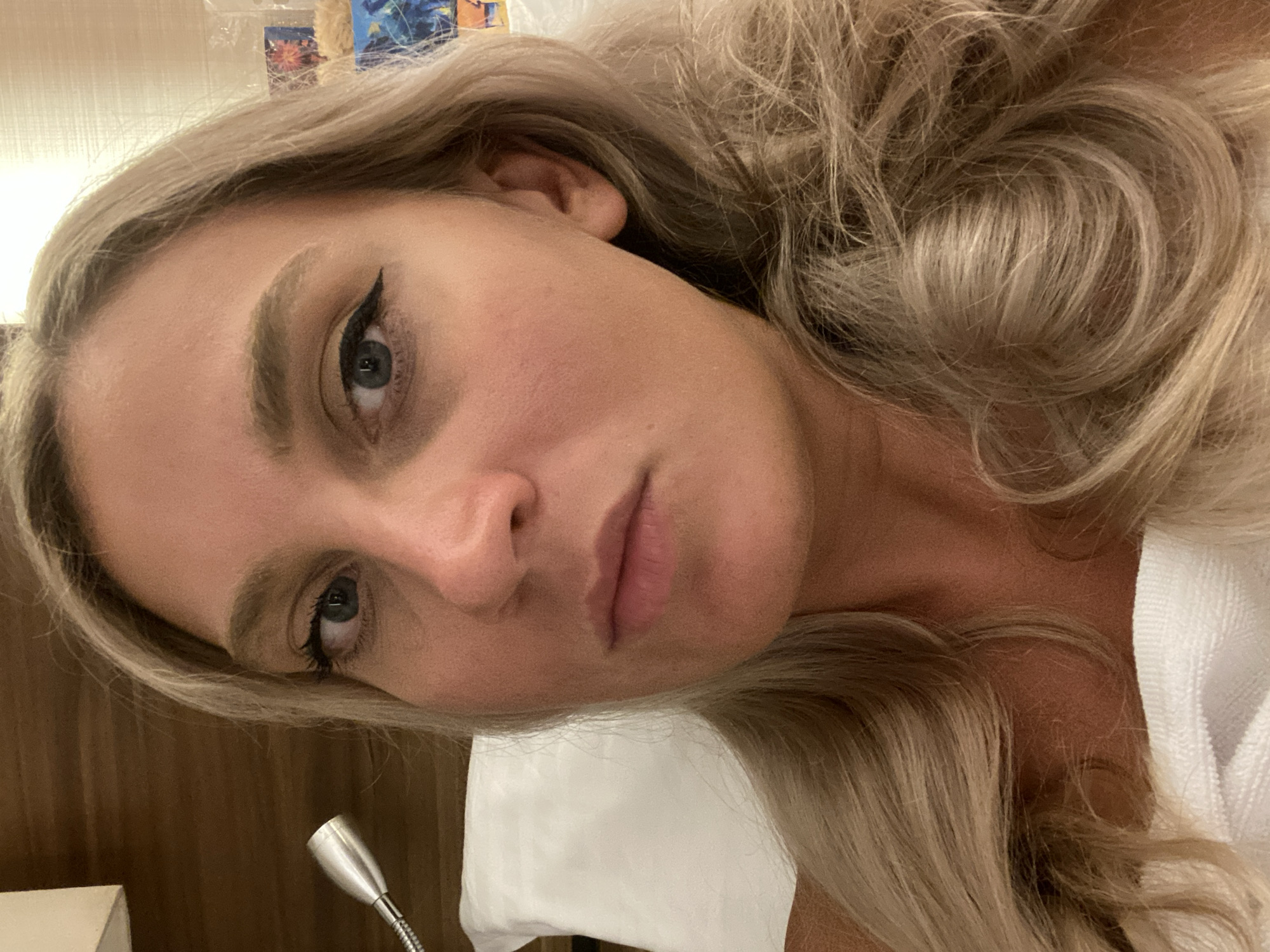 The image is a close-up selfie of a blonde woman in Aksu, Turkey, lying on a bed. She is wearing makeup, including winged eyeliner, and her hair is styled in loose waves. The background shows a headboard of a bed and a wall with a textured pattern. To the left, a silver gooseneck reading lamp is visible.