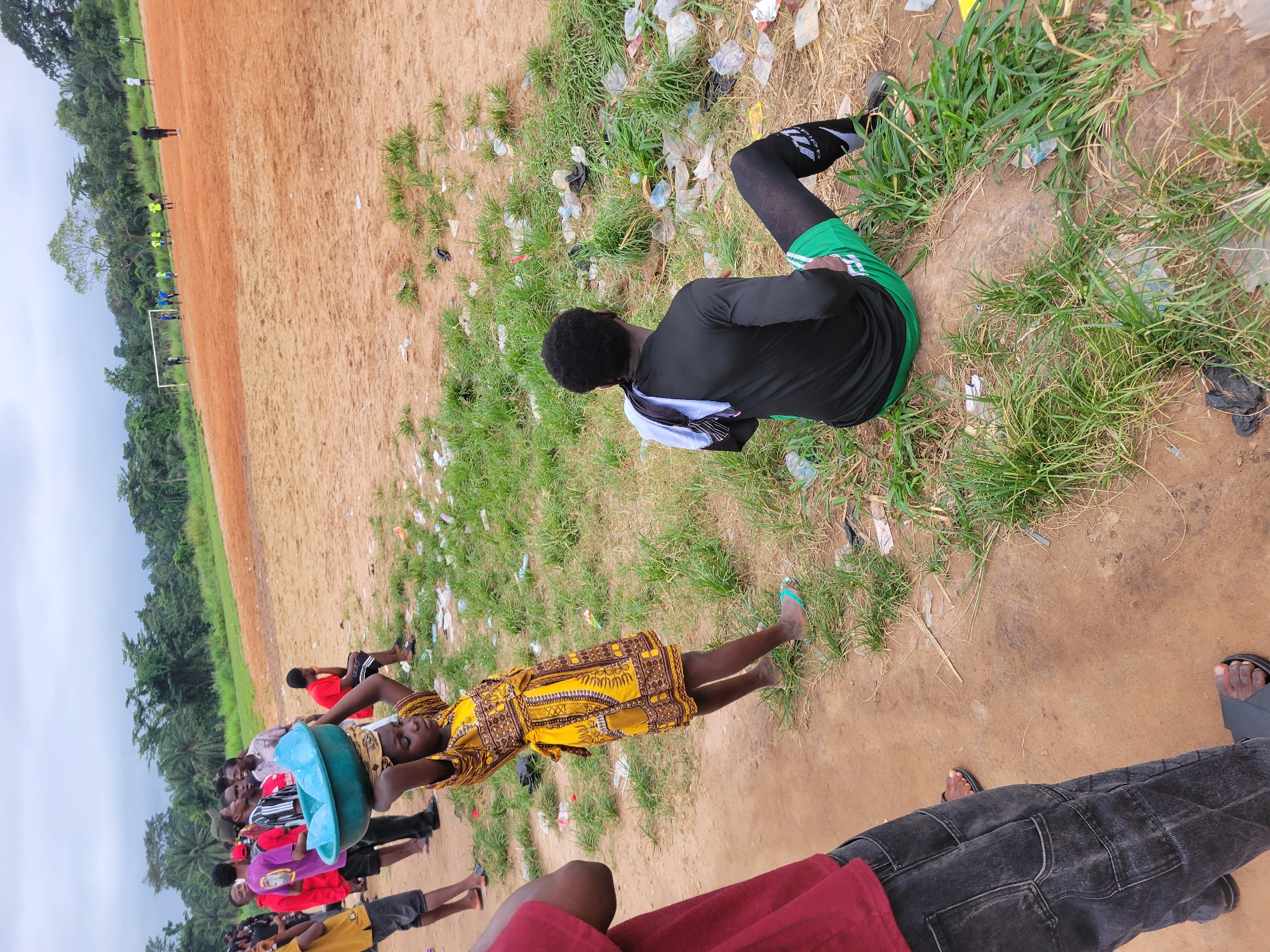 In Egbagu, Nigeria, on a dusty, outdoor football field, a group of individuals are gathered. A young girl, adorned in a yellow and brown patterned dress, carries a teal basin filled with water on her head. She stands near a collection of people dressed in casual attire, some wearing bright red and purple shirts, watching a football game in the distance. A young man, wearing a black long-sleeved shirt, green shorts, and black and white socks, sits on the ground facing away from the camera, a white towel draped over his shoulder. The field is littered with trash, including plastic bottles and wrappers, interspersed with patches of dry grass. The sky above is overcast, and a line of trees borders the field in the background.