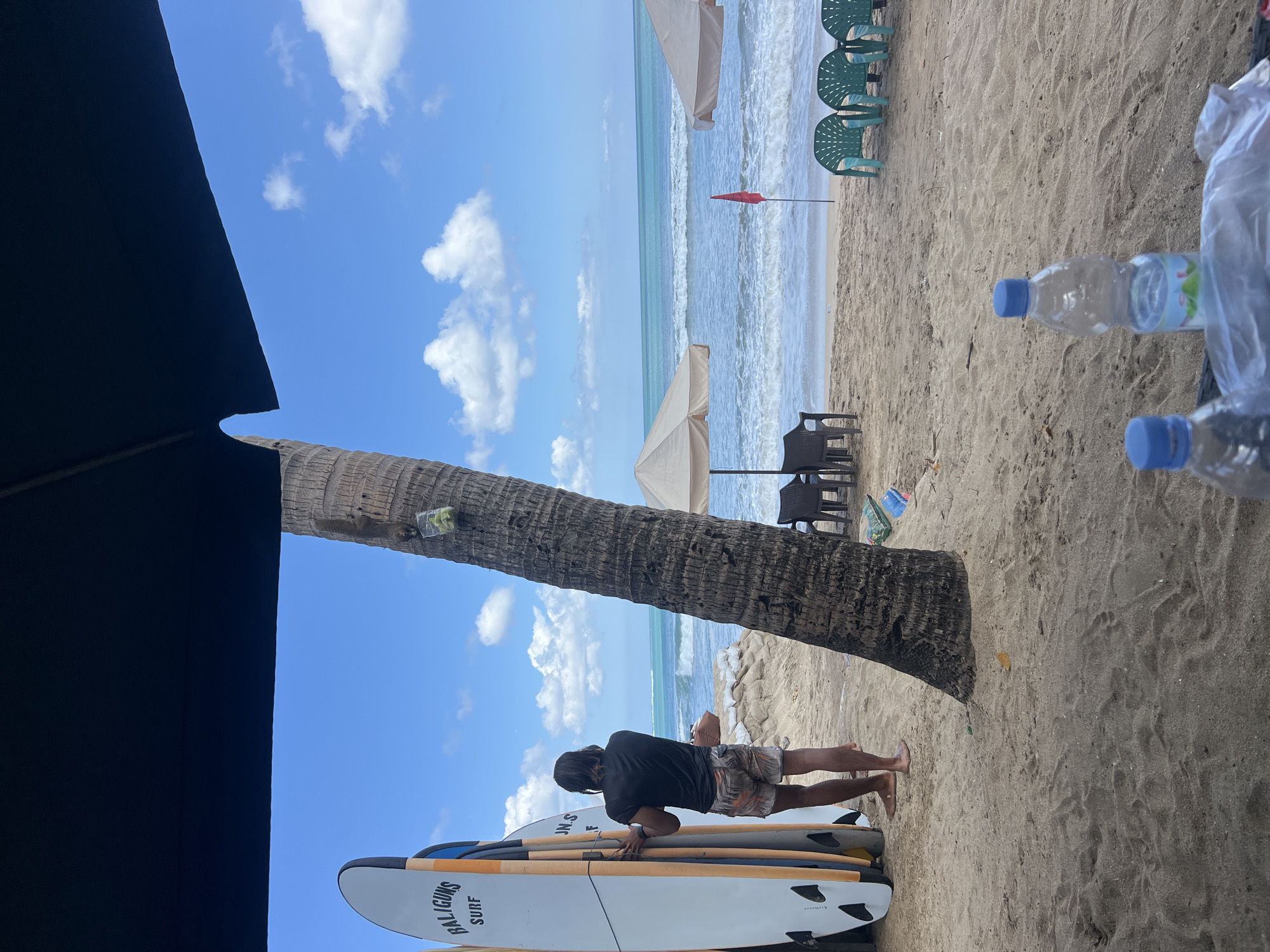 A person stands on a sandy beach in Kuta, Indonesia, facing the ocean. They are wearing a dark t-shirt and patterned shorts, with their back to the camera. To their left, a row of surfboards stands upright, leaning against a black structure that is partially obscured by the top of the frame. A palm tree with a small plastic bag containing what appears to be fruit or vegetables tied to it stands to the right of the person. Further in the background, beach umbrellas and chairs are set up on the sand. The ocean has moderate waves, and the sky is blue with scattered white clouds. The scene appears to be a typical day at a beach resort.