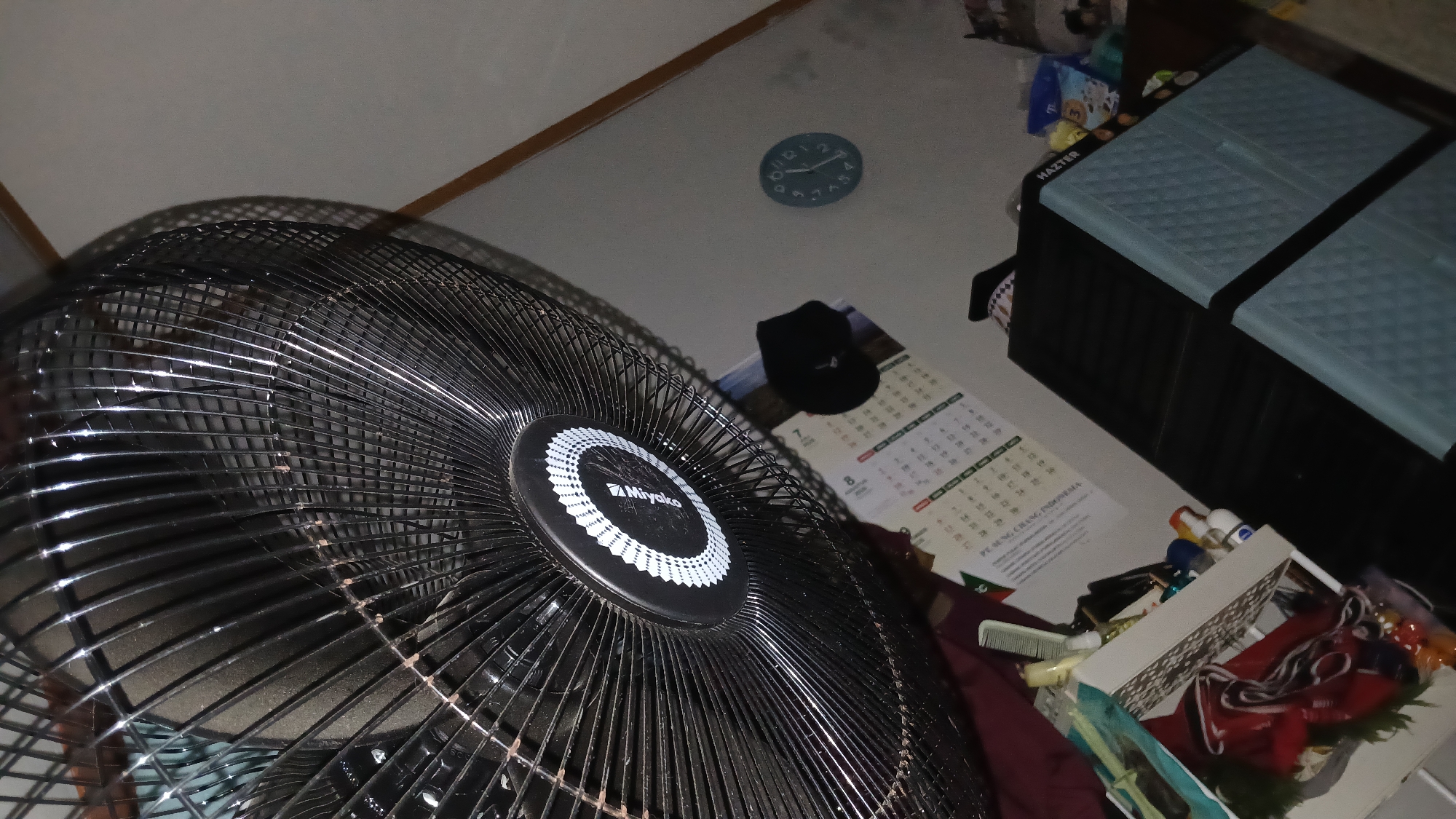 This is a close-up, slightly tilted, indoor shot of a black oscillating fan, positioned in the foreground. The fan's grill and central circular emblem with "Miyako" branding are visible. To the right of the fan, a light blue wall is partially visible, featuring a circular clock with black hands and numerals. Below the clock, a calendar hangs on the wall. In the lower right quadrant of the image, a black storage unit is visible, topped with a light blue quilted lid. Various objects, including bottles and what appears to be a comb, are scattered on a surface to the left of the storage unit. The overall scene suggests a domestic or storage area, likely in Sampang, Indonesia, given the presence of the "PT. SUNG CHANG INDONESIA" text on the calendar.