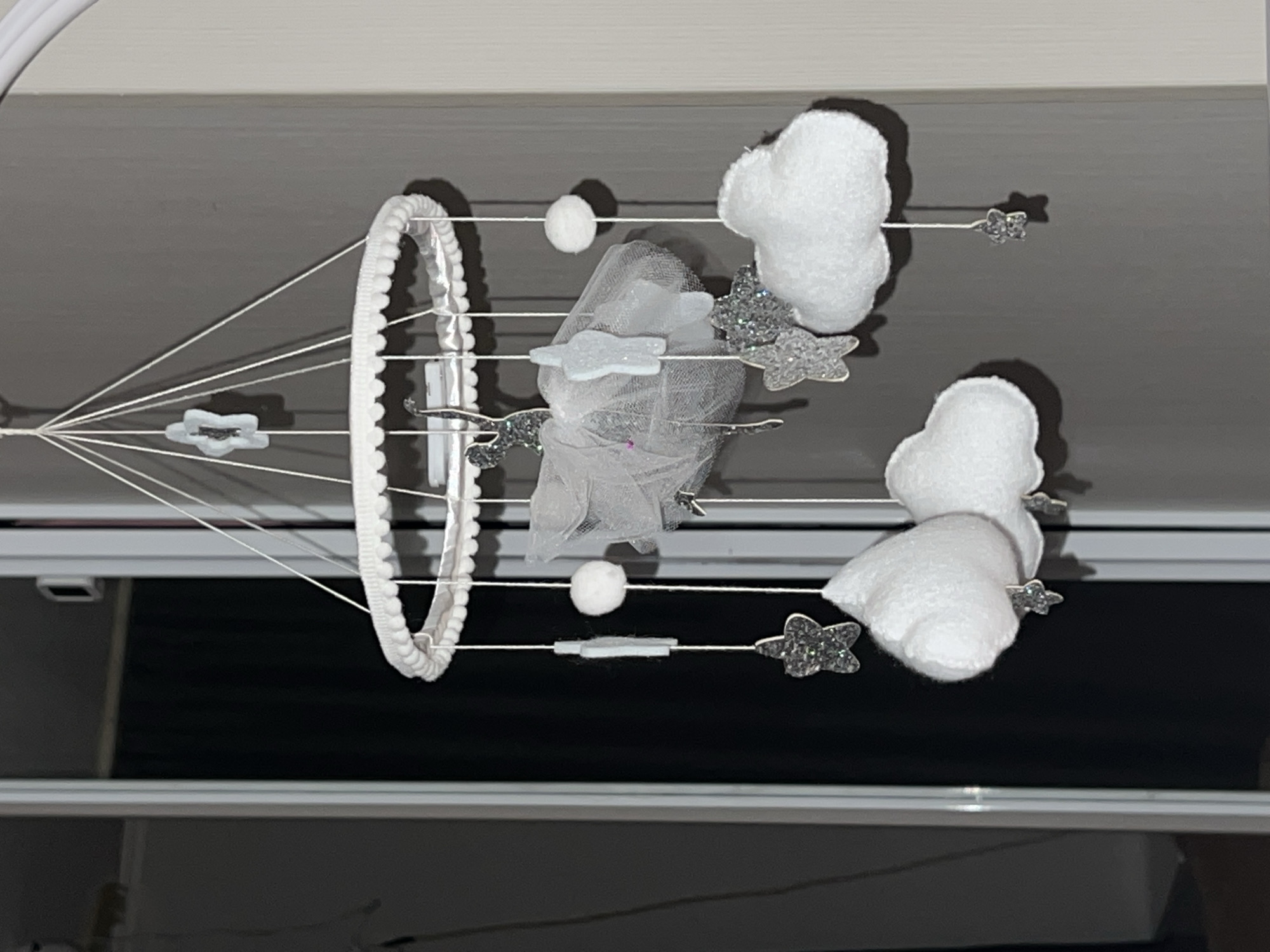 A baby mobile hangs from a ceiling. The mobile features a white beaded ring, white clouds, a white tulle element, silver stars, and white pom-poms. The background is a combination of a ceiling and possibly part of a window frame. There is no indication of the location being Chaikovsky, Russia, or any people or actions in the image.