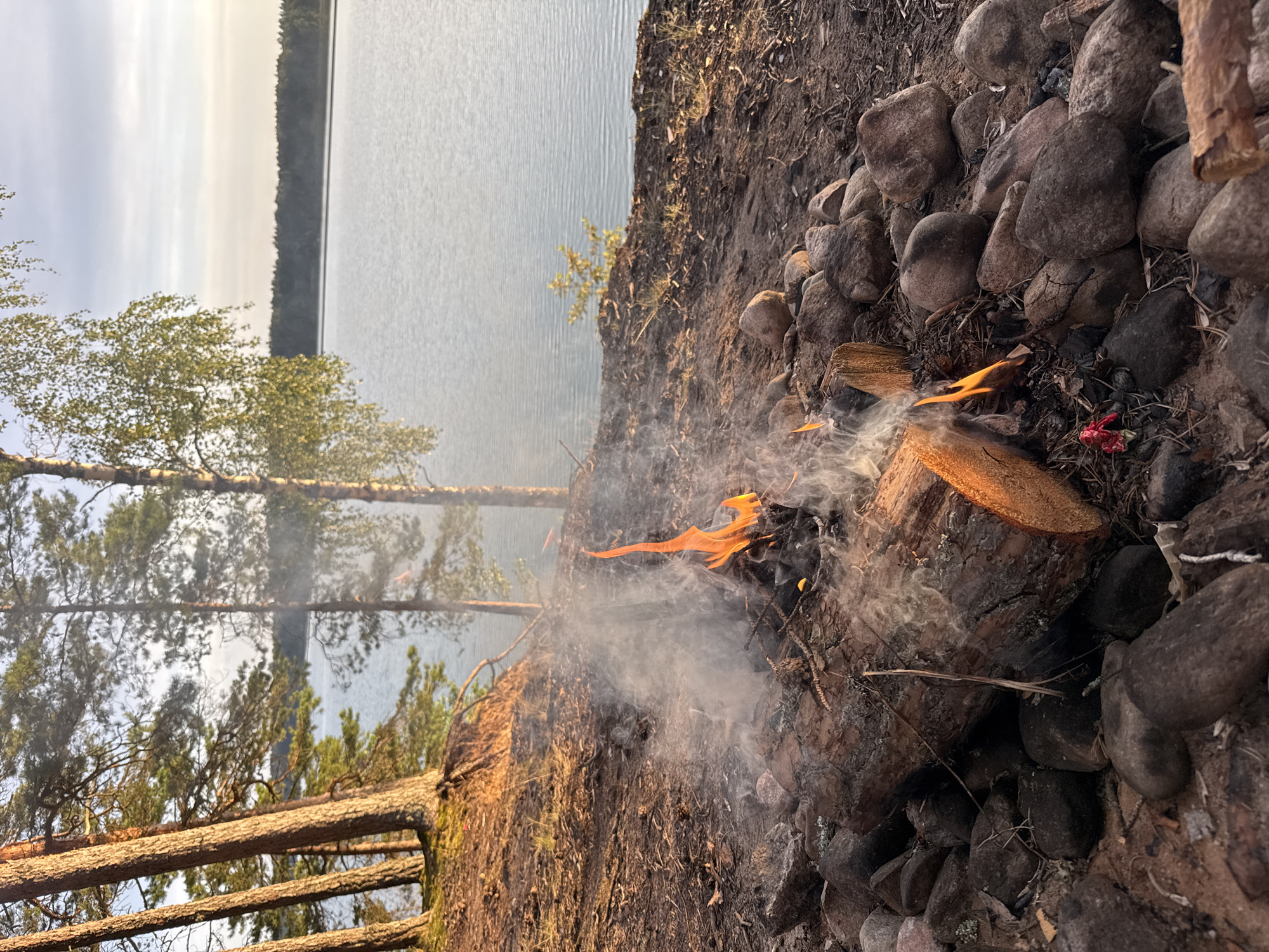 A campfire burns on the rocky shore of a lake, with trees lining the embankment and a forest in the background. The sky is overcast and the water is calm. The scene is tranquil and secluded, likely in a natural area.