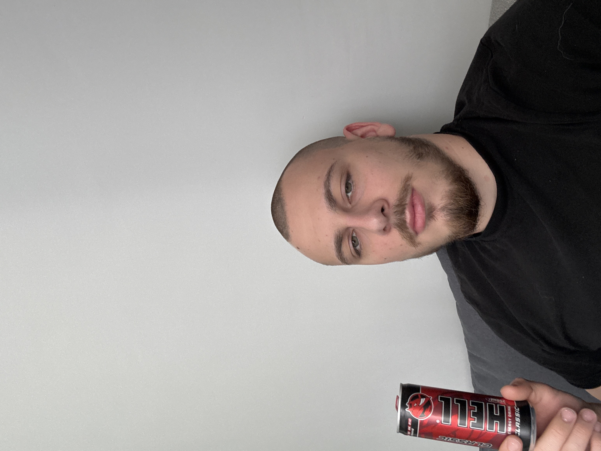 A man with a shaved head and a beard is holding a can of "Hell" energy drink. He is wearing a black t-shirt. The background is a plain white wall. The image is rotated. There is no information provided that allows for the identification of the location in the city of Stara Zagora, Bulgaria.
