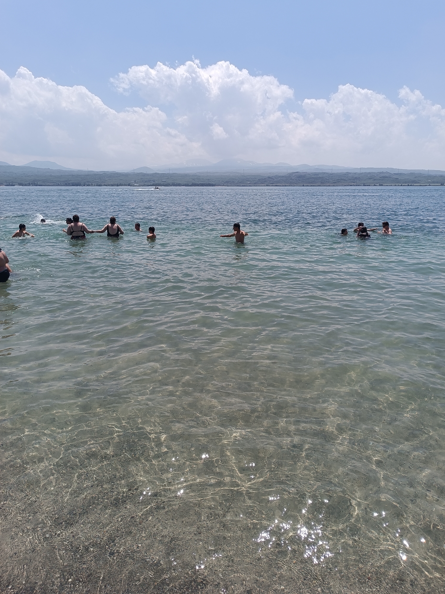 In this medium shot taken on a sunny day, a group of people are enjoying a swim in a clear, calm lake. The water is shallow enough near the shore for the subjects to be standing, with some further out swimming or treading water. Several children and adults are visible, some with their backs to the camera, others facing away. The scene is set against a backdrop of hazy, blue-hued mountains under a bright blue sky dotted with fluffy white clouds. A small boat can be seen in the distance. The overall impression is one of a leisurely, recreational activity on a pleasant day at a lake, likely a popular spot for swimming and cooling off. The clear water reveals the rocky bottom, creating a shimmering effect with the sunlight. The location is identified as Sevan, Armenia, suggesting this is Lake Sevan.