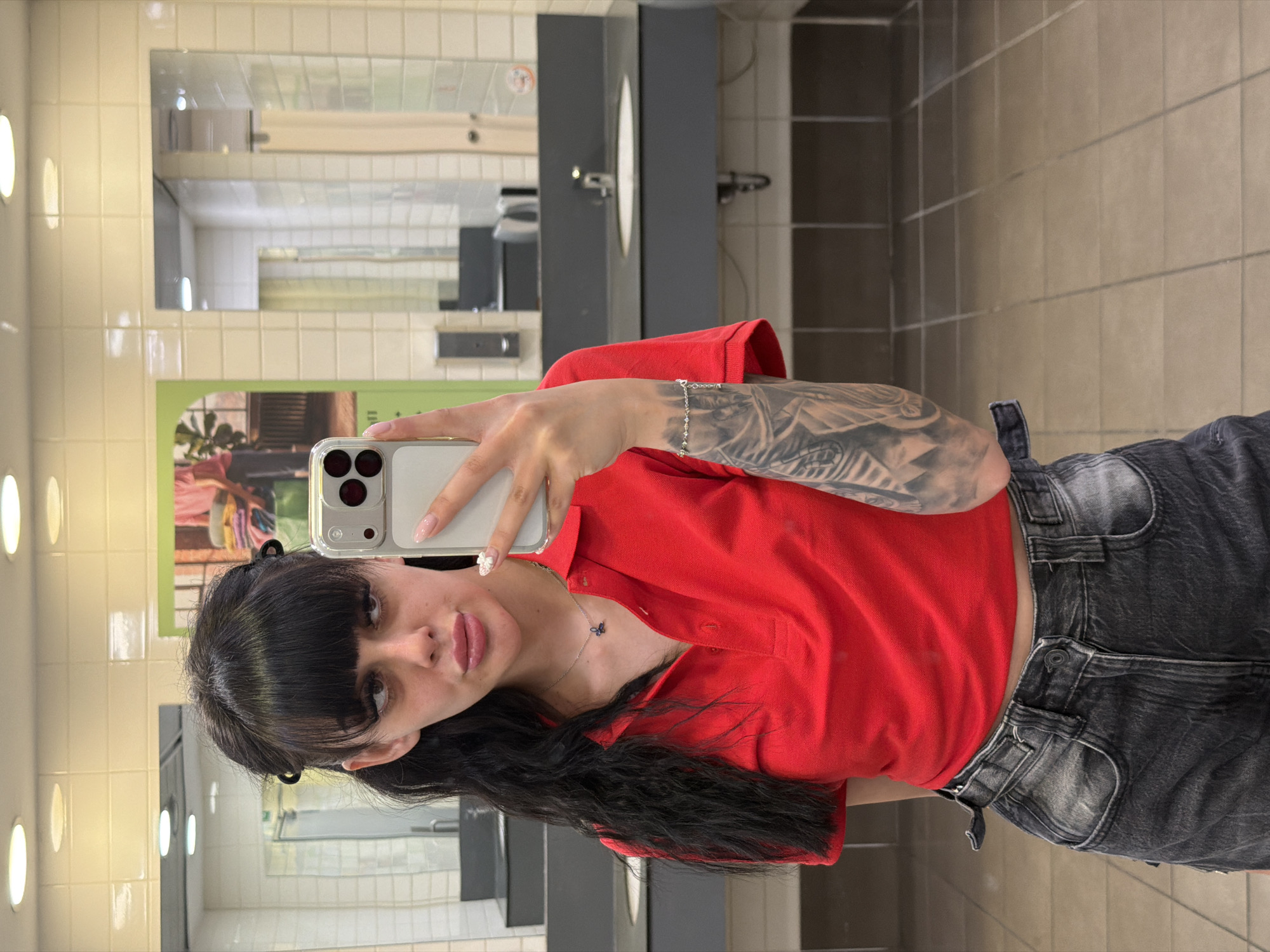 A young woman with dark hair and bangs is taking a mirror selfie in a public restroom. She is wearing a red polo shirt and dark gray denim shorts. She has a tattoo on her left arm and is wearing a silver necklace with a blue butterfly pendant and a silver bracelet. The restroom has tiled walls and floors, a gray countertop with sinks, and several mirrors. A framed picture is visible on the wall behind her. The image appears to be taken in a well-lit, modern restroom facility, potentially in a public building, shopping center, or service area within the city of Plovdiv, Bulgaria.