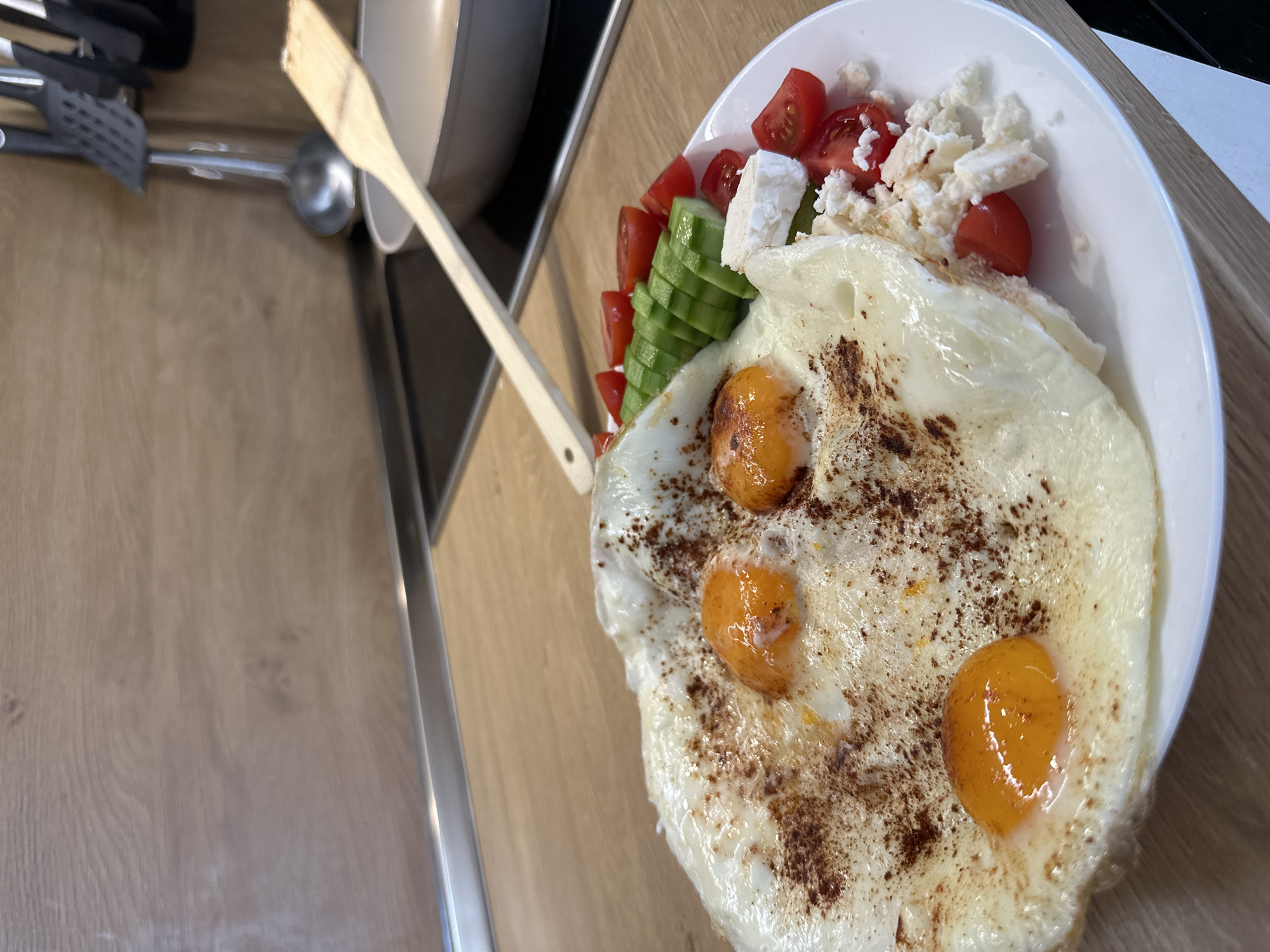 The image captures a close-up, overhead view of a freshly prepared meal, likely breakfast or brunch, resting on a light-colored wooden kitchen countertop. The location context is Plovdiv, Bulgaria. The main subject is a white, oval-shaped plate filled with food. Dominating the plate are several fried eggs, distinguished by their separate, bright orange yolks and mostly set white albumen. A dark reddish-brown spice, possibly paprika or sumac, is sprinkled liberally over the eggs. Adjacent to the eggs is a vibrant salad component, consisting of neatly diced red tomatoes, thinly sliced green cucumbers arranged in a row, and generous crumbles of white cheese, likely feta, scattered among the vegetables.
In the immediate background, a wooden spatula or spoon rests with its handle extending towards the left side of the frame. Further back, out of focus, other kitchen utensils such as a metal ladle and black spatulas are visible, alongside a portion of a kitchen appliance, possibly a blender or coffee machine. The wooden countertop features a visible grain pattern and has a metallic strip running along its edge, suggesting a modern kitchen setting. No people are visible, and there is no discernible text. The scene depicts a meal ready to be consumed, suggesting a morning or early afternoon setting.