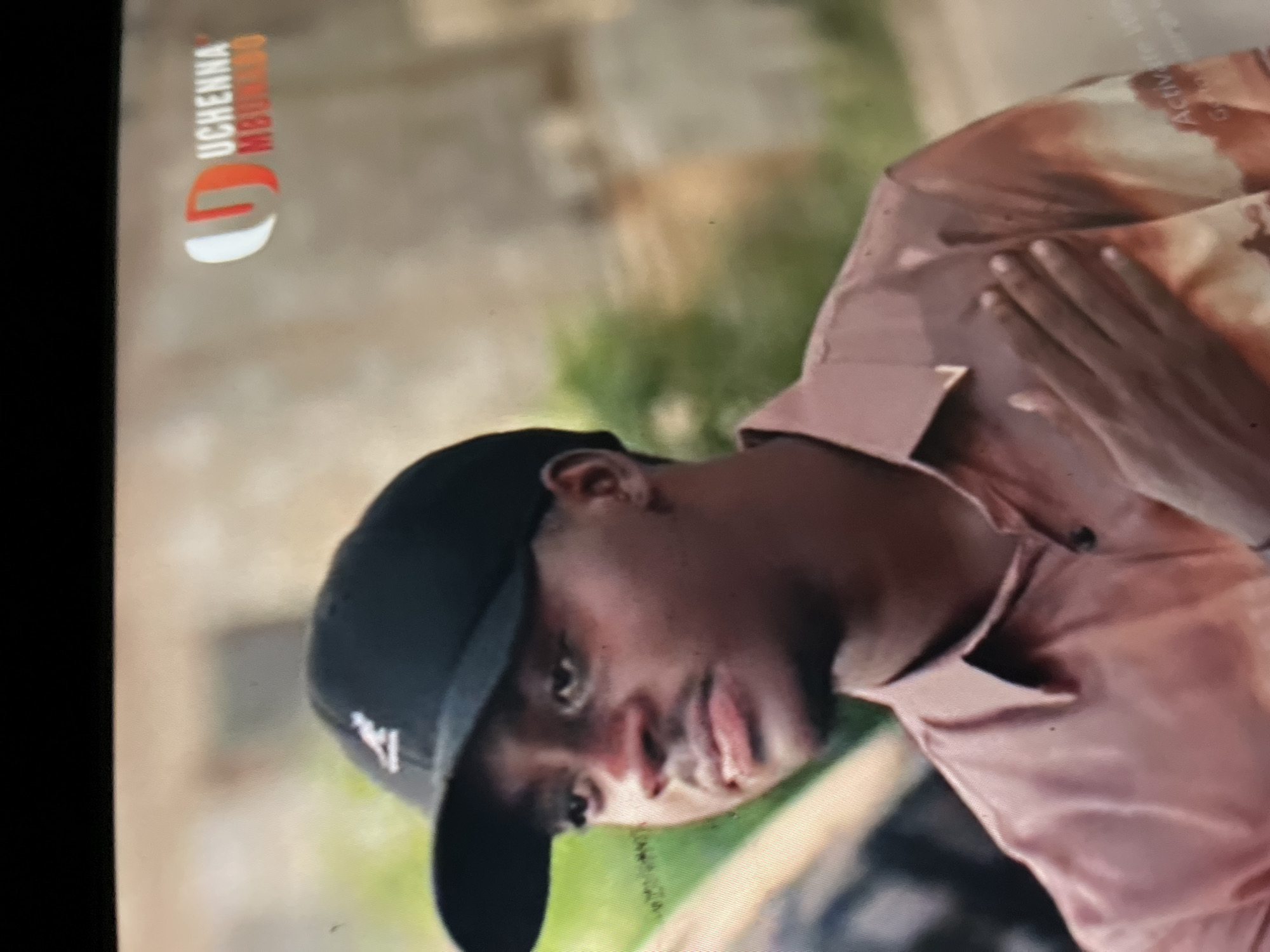 The image features a young man of African descent, captured from the chest up, in what appears to be an outdoor setting in Abuja, Nigeria. He is looking directly at the viewer with a calm, slightly smirking expression. He wears a dark, possibly black, baseball cap with a distinct white logo on the side, resembling a stylized 'W' or 'V'.
The man is dressed in a light-colored, possibly rose-gold or beige, collared shirt that exhibits a subtle, abstract, or tie-dye pattern in varying shades of brown and tan. The fabric has a soft, slightly lustrous appearance. His right hand rests gently on his left shoulder or upper chest, with fingers spread.
The background is blurred, indicating a shallow depth of field, and shows hints of green foliage and lighter, earthy tones, consistent with a daytime outdoor environment. The lighting is bright and even.
In the top left corner of the image, there's a prominent graphic overlay displaying "UCHENNA MBUNDU" in white and orange/red text, positioned above a red and white logo that forms a stylized 'U' or 'G'. Faint, partially illegible text, possibly "Activ" and "Crea", is also visible in the upper right section of the image. The entire image is noticeably tilted.