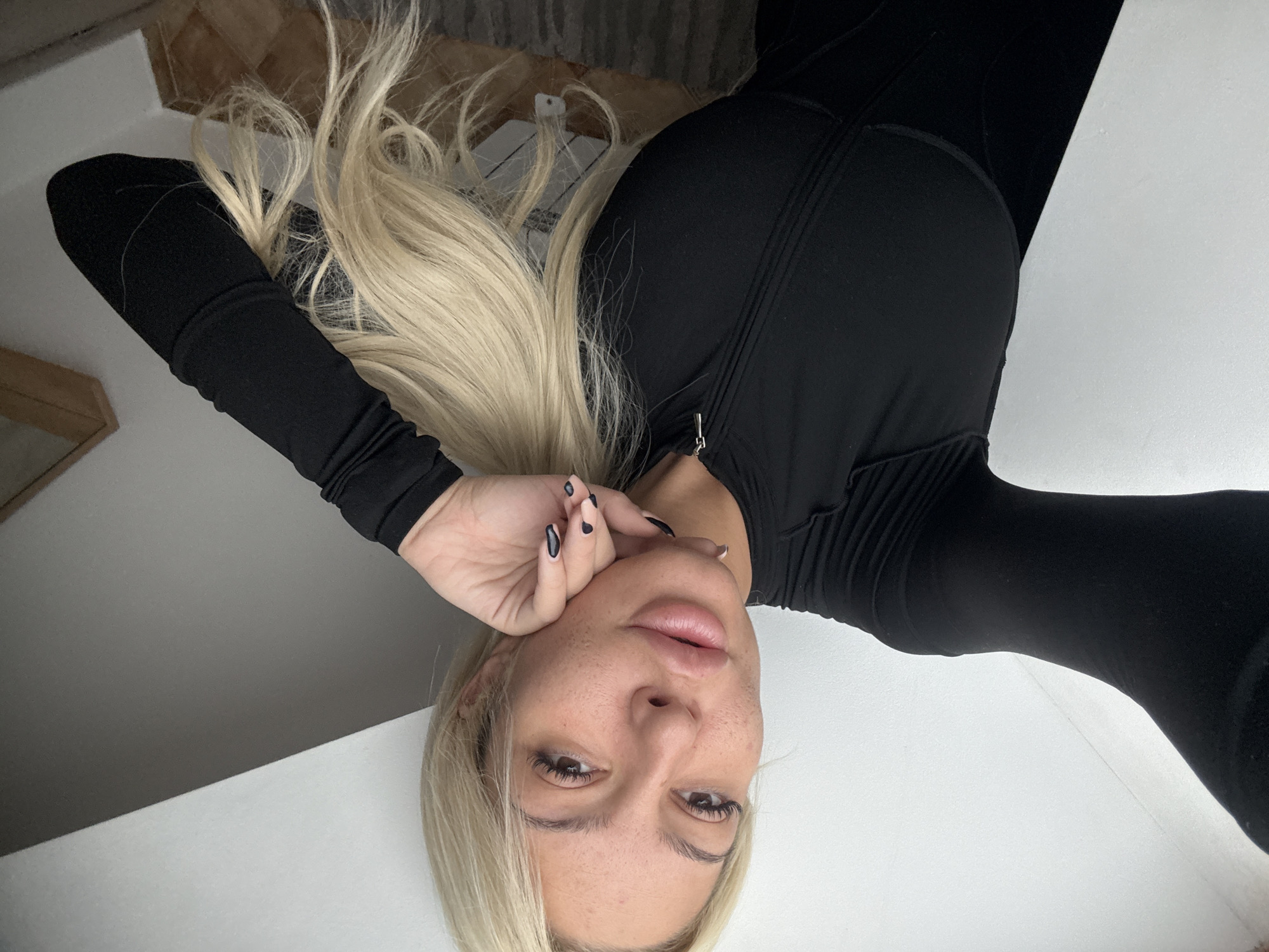 An indoor, high-angle photograph taken in Sofia, Bulgaria, features a person, likely female, with long, flowing blonde hair. The image is composed with the subject appearing upside down, looking directly into the camera while seemingly lying or reclining on a surface.
The subject is wearing a form-fitting black long-sleeved top, which appears to have a zipper closure running down the front or side of the torso. Her blonde hair is spread out around her head and upper body. Her face displays full lips, prominent dark eyelashes, and visible freckles across her nose and cheeks. Her expression is neutral and direct.
Her right hand is visible, resting against her face, showcasing manicured nails with a distinctive two-tone design, featuring a light base and black tips or pattern. The background is predominantly a clean, bright white or light-colored surface, possibly a bed or floor. Hints of darker, textured elements are visible in the upper corners, suggesting other indoor furnishings or walls. The lighting is bright and even, illuminating the subject clearly. No visible text is present in the image.