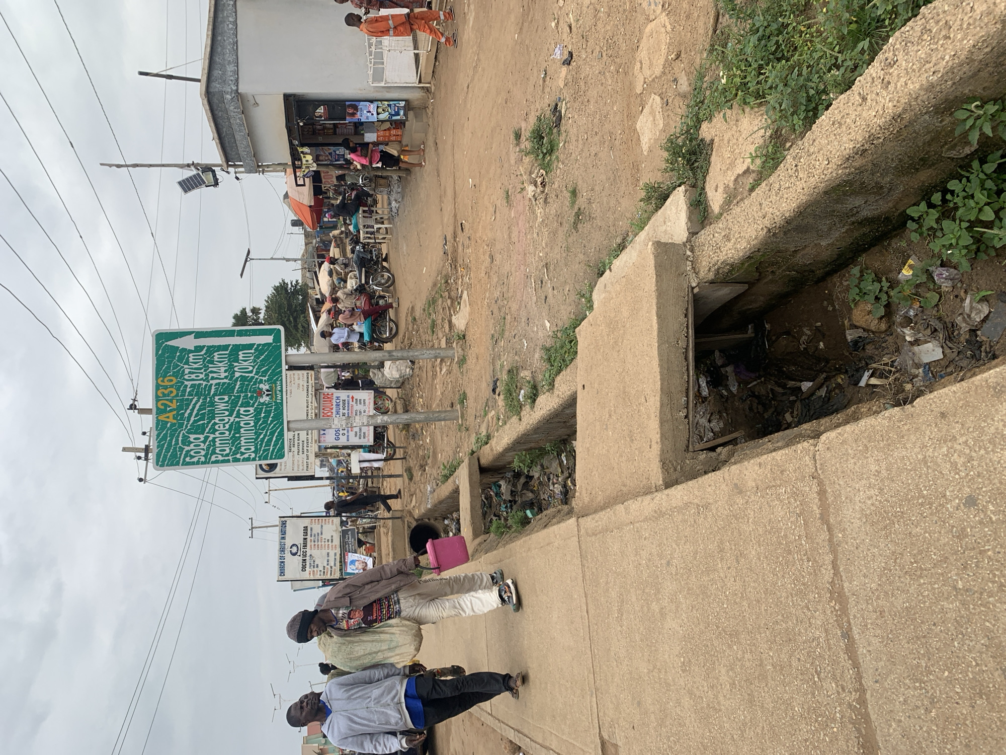 The image shows a street in Rafin Par, Nigeria. A concrete-lined drainage ditch runs along the edge of the street, filled with trash. Several people are walking on the sidewalk, including two men carrying items, one with a pink bag. A sign indicates "A236" with distances to Sobd (87 km), Pambeguwa (144 km), and Saminaka (10 km). Several signs advertising churches are visible. The street is unpaved and leads to a small market area with shops and stalls. Motorbikes are parked in the market area. Utility poles and wires are overhead. The sky is overcast.