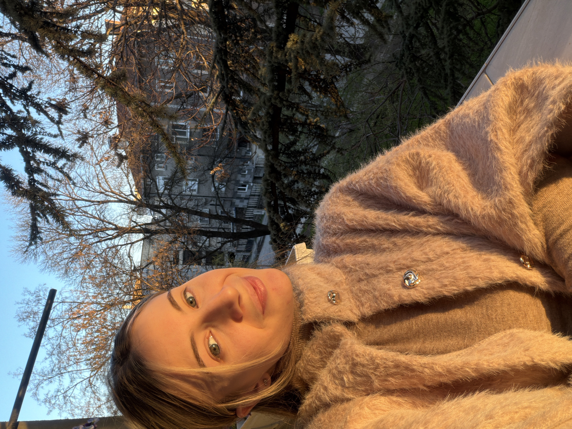 A woman is pictured outside in an urban setting, likely in Plovdiv, Bulgaria, based on architectural style and vegetation. The woman is fair-skinned with blonde hair and green eyes. She is wearing a light brown sweater and a fuzzy, buttoned cardigan. Behind her is a building with multiple windows and a tree with sparse branches. The sky is blue.