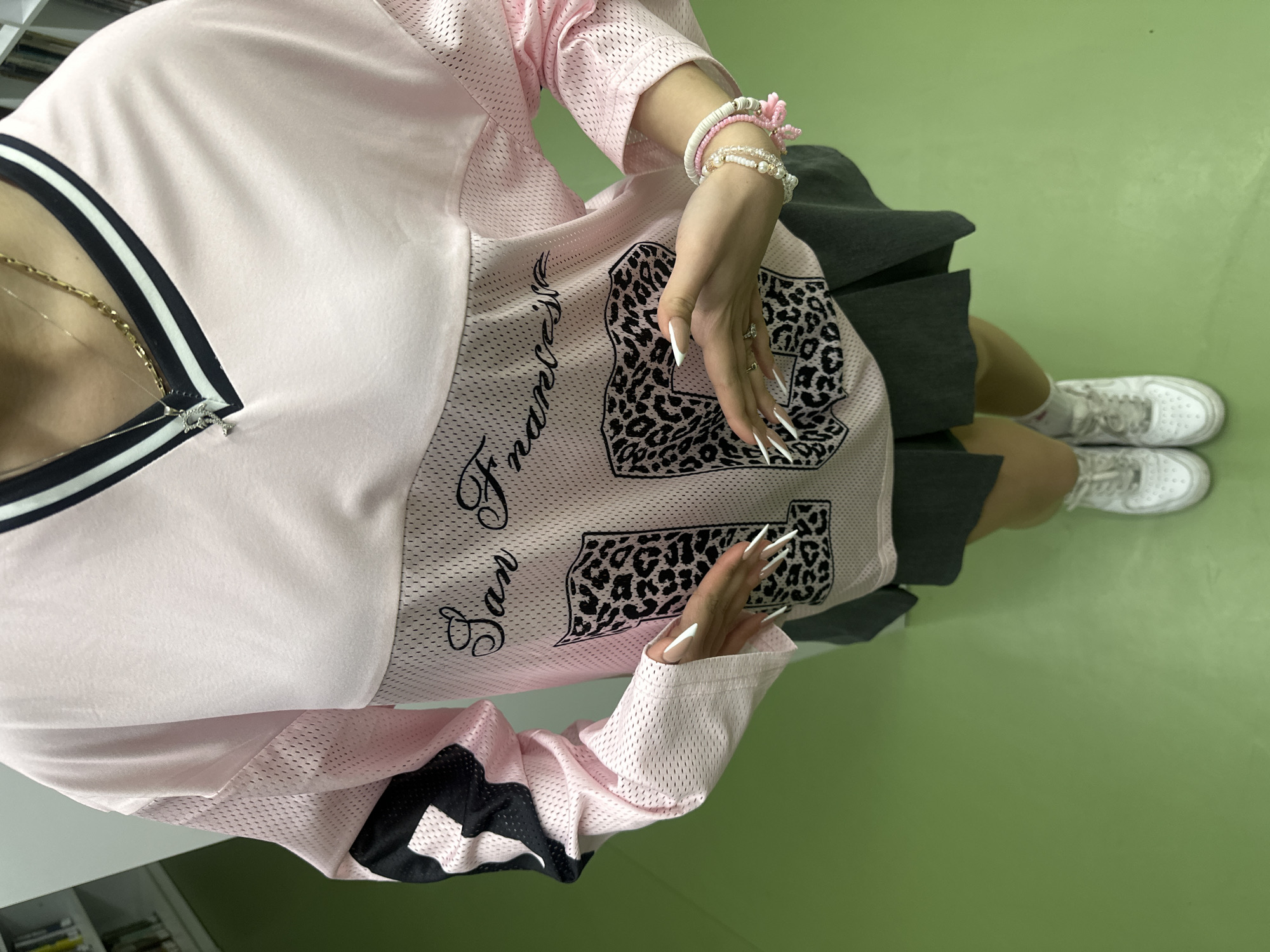 A partial, downward-angle image depicts an individual, likely female, standing indoors in Yerevan, Armenia. The subject is wearing a light pink mesh athletic jersey with a V-neck collar featuring black and white stripes. The jersey has "San Francisco" in black cursive script and large, leopard-print numbers, primarily "15," visible on the front. A portion of a black and white geometric pattern is on the upper left sleeve. The individual wears a dark gray pleated mini skirt, white socks, and white athletic sneakers, likely Nike Air Force 1s. Two necklaces are visible: a thin silver chain with a small, sparkling pendant (possibly an initial 'P' or 'R') and a thicker gold chain. The right wrist displays multiple bracelets, including strands of white and pink beads/discs. Both hands are held up at waist level, palms open and facing each other, with fingers slightly spread. All visible fingernails are long, pointed, and white. The immediate environment consists of a solid light green floor. In the blurred background, a light-colored shelving unit with various items is partially visible.