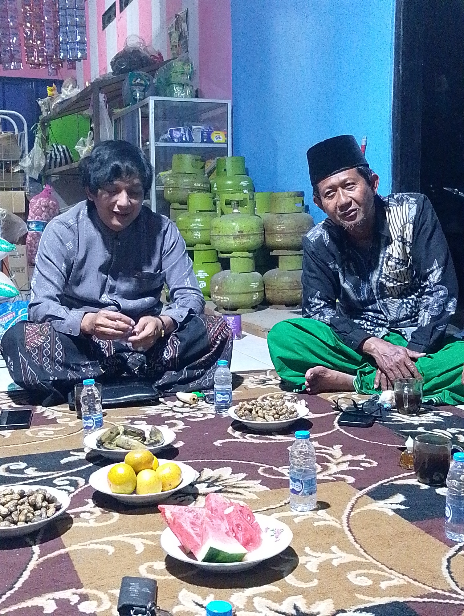 Two men are sitting on the floor in a dimly lit room, presumably in Cilacap, Indonesia. They are both dressed in traditional attire. One man on the left wears a grey patterned shirt and dark patterned pants, while the man on the right is wearing a black and white patterned shirt and green pants, with a black peci (cap) on his head. They are facing each other, with food and drinks spread out on a rug between them. The food includes peanuts, bananas, oranges, and watermelon. Several bottles of water and glasses of a dark beverage are also visible. In the background, stacks of green gas cylinders are prominent, along with shelves holding various items. The overall atmosphere appears to be casual and social.