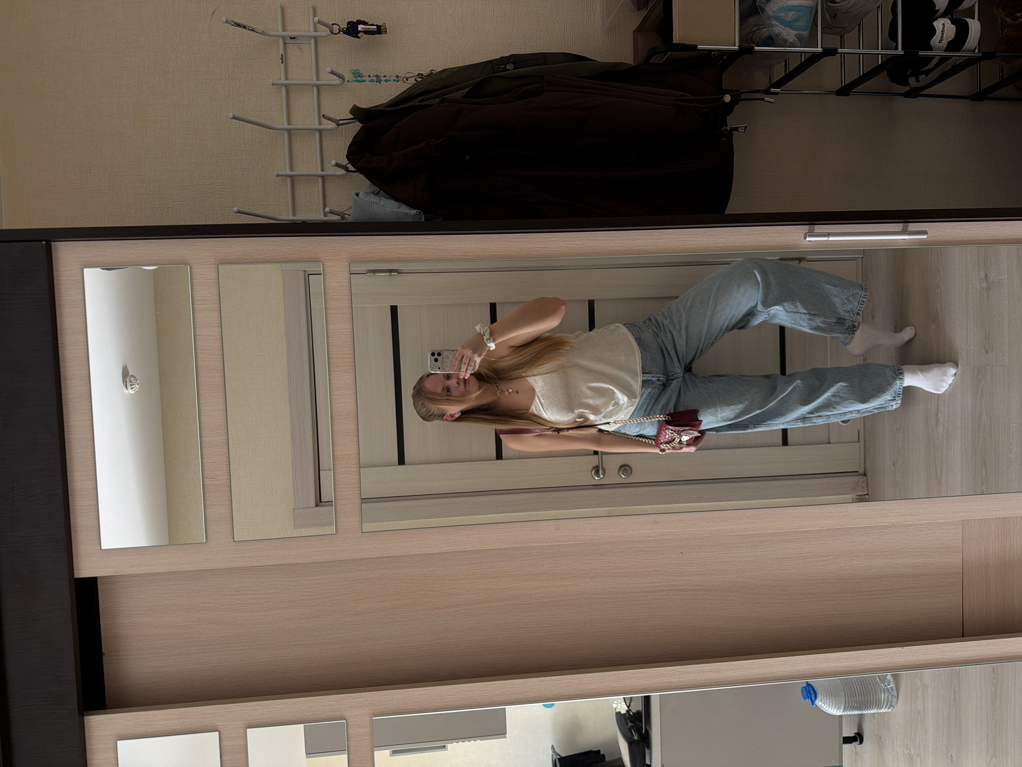 A young woman is taking a selfie in a mirror located on a light-colored wooden wardrobe. She is wearing a white top, light blue jeans, and white socks, and is holding a cell phone in her left hand. She has long blonde hair. A small red purse hangs from her right shoulder. The mirror reflects a partially visible interior, including a wooden door with vertical panels. Above the wardrobe, hanging on a wall, are a coat rack with jackets, and a shoe rack with shoes. Below the wardrobe in the mirror, is a water bottle. The location is implied to be in Izhevsk, Russia, though there is no visible information to confirm this. The primary action is the woman taking a selfie.