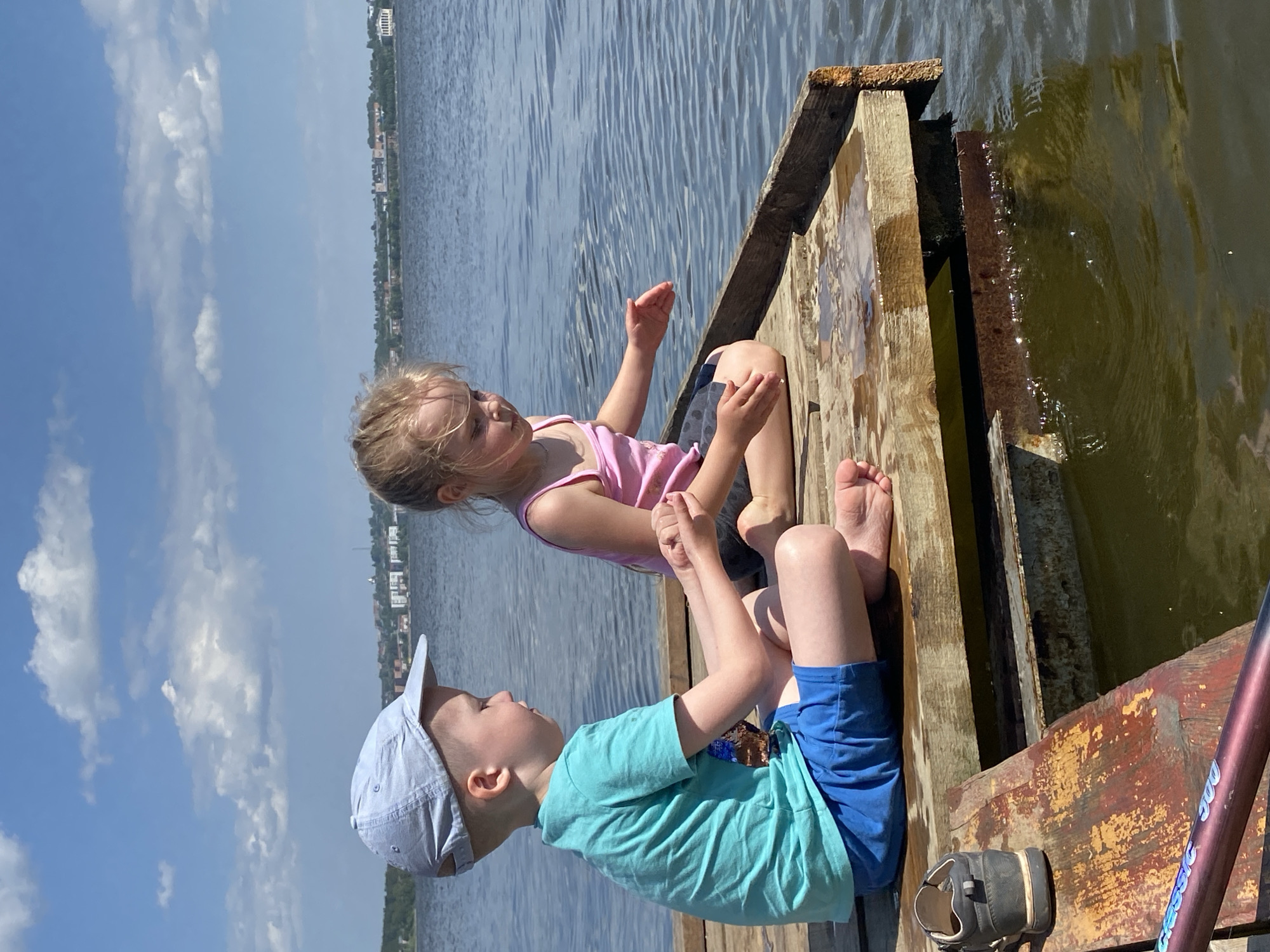 Two young children, a boy and a girl, are sitting on a weathered wooden dock at a waterfront in Votkinsk, Russia. The girl wears a pink tank top and dark shorts. The boy wears a light blue t-shirt, blue shorts, and a light blue baseball cap. He is looking up at the girl, and they appear to be interacting. To the right of the children, partially submerged, is the dock’s structure; water is visible. In the background is a body of water. Across the water, the cityscape of Votkinsk is visible under a blue sky with scattered clouds. A shoe sits on the dock. A boat paddle lies on the far right edge of the frame.