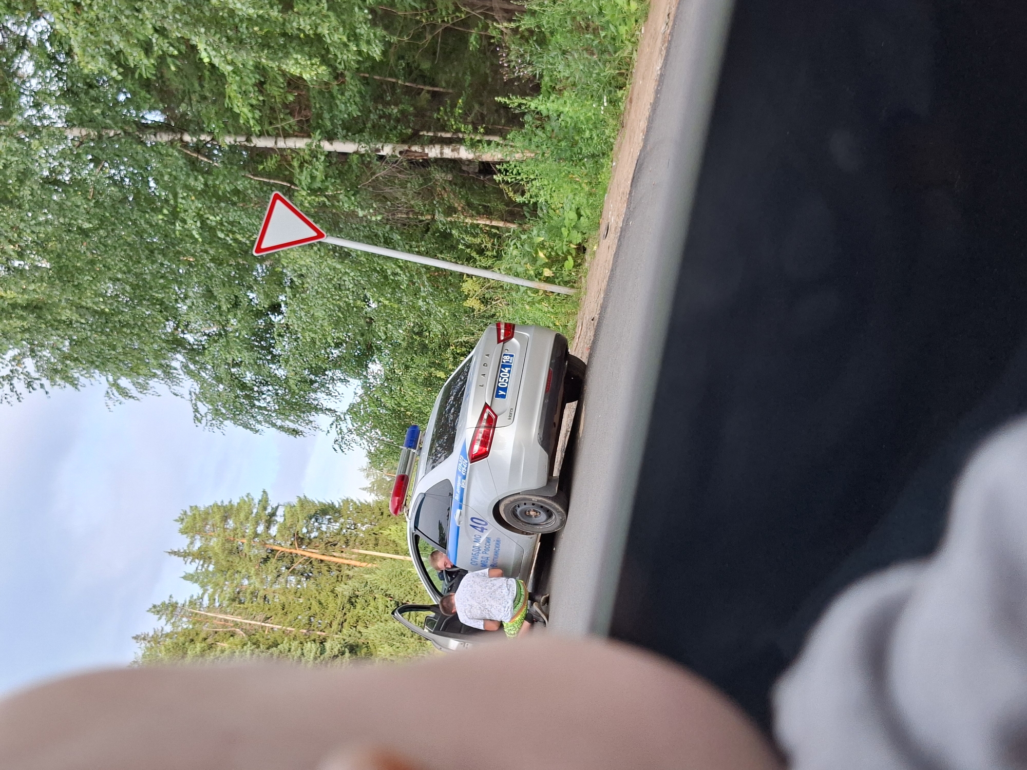 In an outdoor, rural or suburban setting with dense green trees lining the right side of a paved road, a silver LADA police car with flashing blue and red lights is parked on the right shoulder. A young man wearing a white t-shirt and patterned shorts is crouched down on the left side of the police car, near the open driver's side door, facing the driver. The driver of the police car, also a young man, is partially visible inside the vehicle. Text on the side of the police car indicates it is affiliated with the "ГИБДД МО МВД России", which translates to "Traffic Police of the Ministry of Internal Affairs of Russia". The text below that appears to be "Воткинский", suggesting a connection to the city of Votkinsk, Russia. A triangular "Yield" sign is visible on a post to the right of the police car, amidst the foliage. The foreground of the image is dominated by a blurred view from inside a vehicle, showing a dark surface in the lower portion and a light-colored, out-of-focus object on the left, possibly a person's arm or shoulder. The sky above is a light blue with some clouds.
