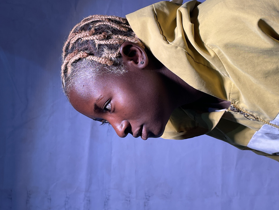 The image displays an individual, with dark skin, positioned in a profile view facing to the left against a textured, light blue background. The subject's hair is styled in numerous small, light-blonde braids/cornrows, with darker roots visible at the scalp. A small, silver-toned stud piercing is present on the left nostril, and a small, dark stud earring is visible in the left earlobe. The eyes are open and directed upwards and to the left. The lips are closed. The individual is wearing a yellow hooded jacket or hoodie; the hood is up and frames the head. A metallic, gold-colored zipper pull is visible on the garment's front, with a segment of white fabric appearing below the yellow on the garment's right side. This scene is situated in Barakin Akawo, Nigeria.