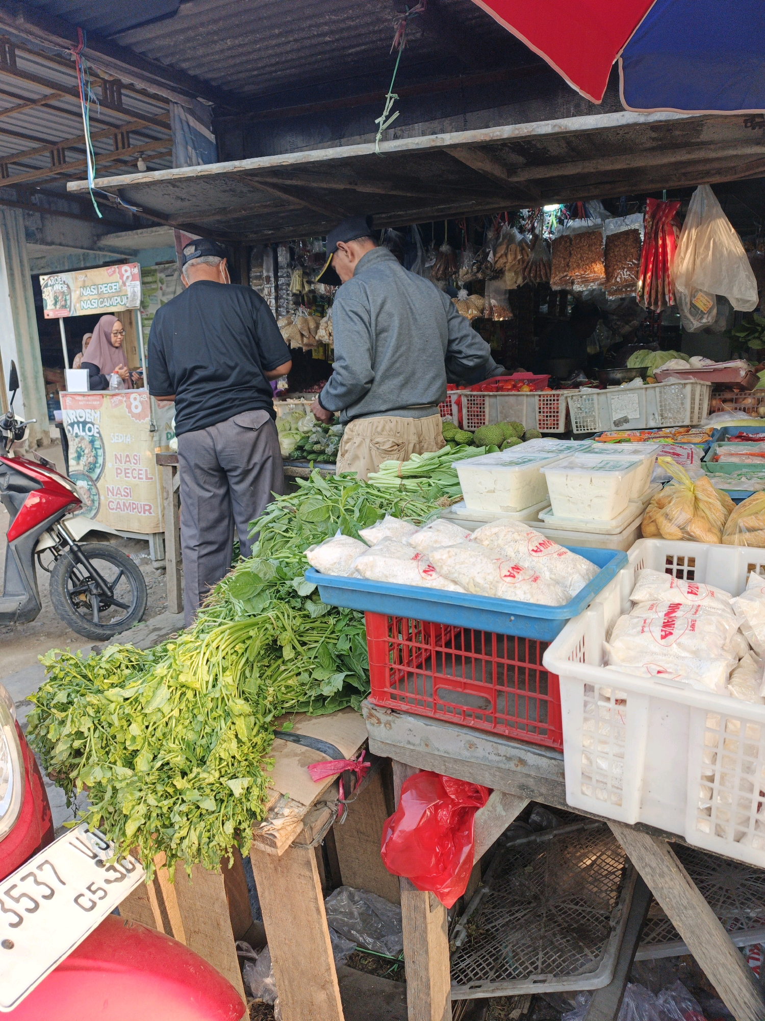 The image depicts an outdoor market scene in Kludan, Indonesia, likely a roadside stall. Two men, dressed in casual clothing and caps, are engaged in activity behind a stall laden with fresh produce and packaged goods. One man, wearing a black t-shirt and dark pants, stands facing away from the camera, appearing to be attending to vegetables. The other man, in a grey long-sleeved shirt and beige pants, is also facing away, seemingly interacting with the vendor or sorting goods. In the background, a woman wearing a hijab is visible behind a counter, with a sign displaying food items like "Nasi Pecel" and "Nasi Campur."
The stall itself is covered by a corrugated metal roof supported by wooden beams. Various items are displayed: bunches of leafy green vegetables are piled in the foreground, while white plastic crates contain what appear to be packaged food items, possibly tempeh or tofu. Other items, such as dried goods in plastic bags and what might be snacks, hang from the stall's structure. A red motorcycle is parked to the left of the scene, its license plate clearly visible. The ground is paved and appears somewhat unkempt, with some debris and plastic bags scattered around. The overall lighting suggests it is daytime, with sunlight casting shadows.