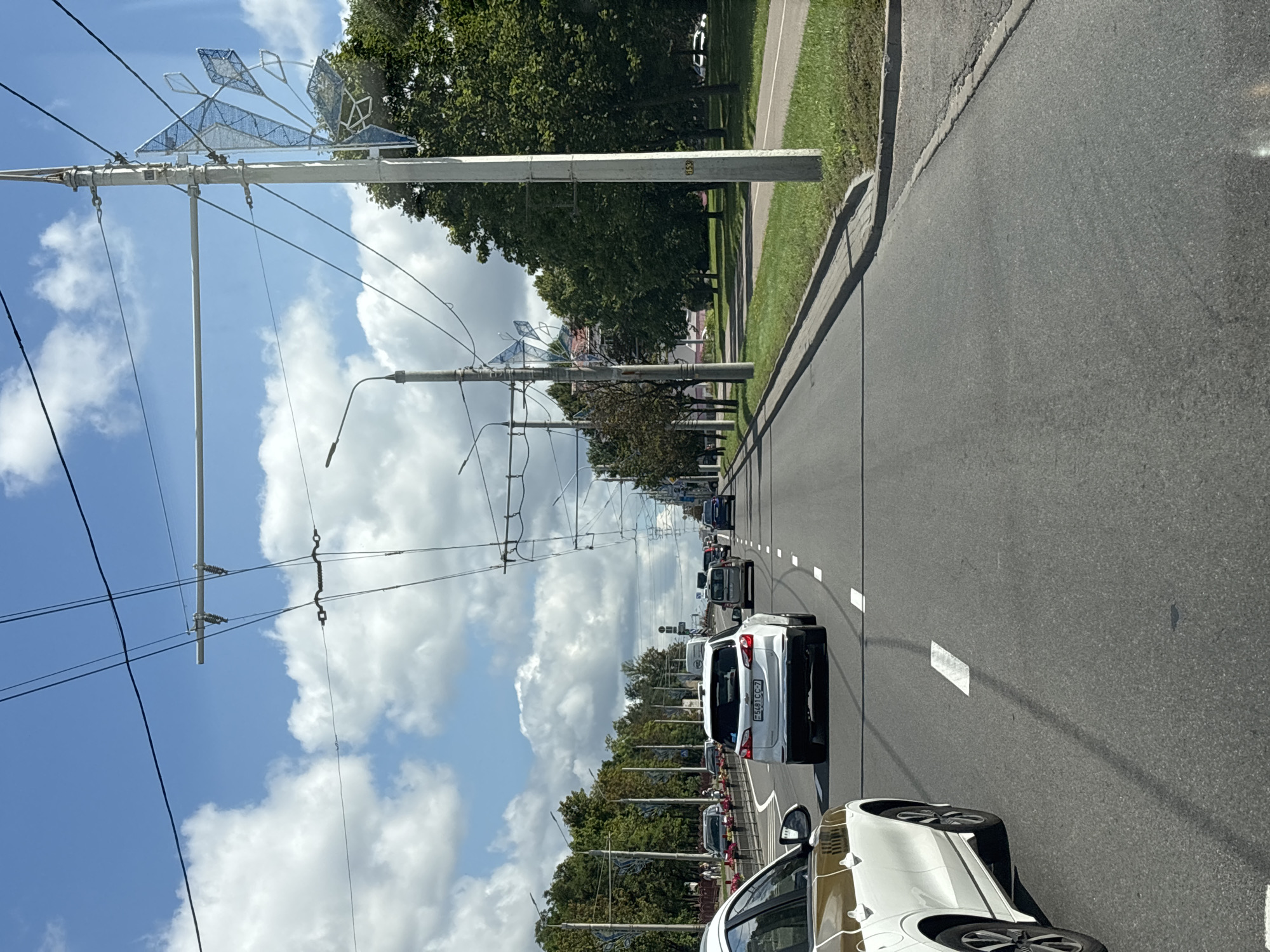 This is a street-level, eye-level, vertical, medium shot of a road in Minsk, Belarus, on a sunny day with scattered clouds. The perspective is from within a vehicle, looking forward. The road has multiple lanes, with white lane markings visible. Several cars are driving on the road, including a white SUV in the foreground with a Belarusian license plate that reads "5481 CC - 7". The road is lined with trees on both sides, and there are trolleybus wires overhead supported by poles. One pole on the right side of the road has a decorative, snowflake-like structure attached to it. On the left side, there is a sidewalk and some pedestrians. The overall scene depicts a typical urban traffic environment.
