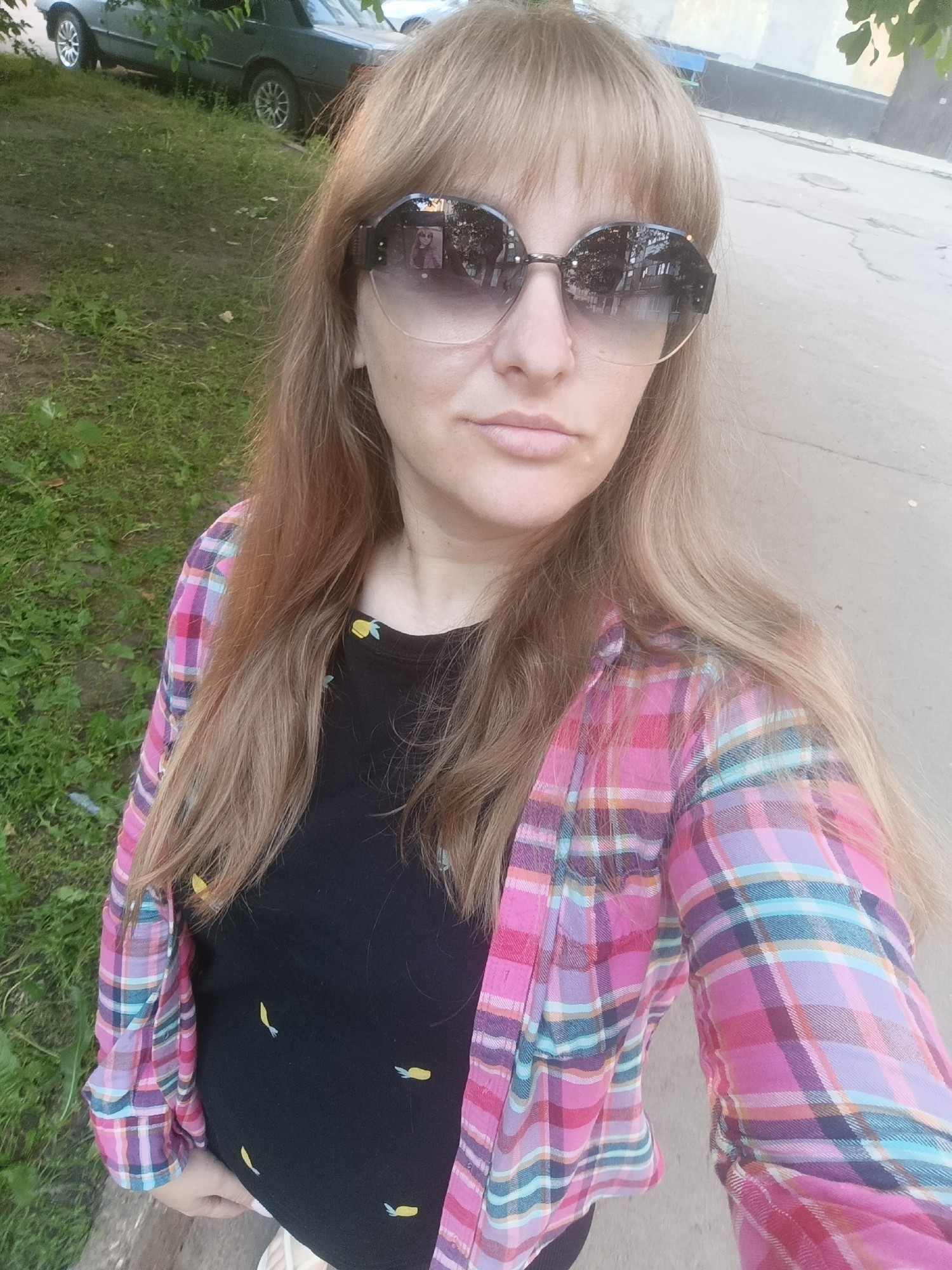 A woman with long, blonde hair and bangs is captured in a selfie. She is wearing large, dark, oval-shaped sunglasses with a subtle gradient tint and a pink, purple, and blue plaid shirt unbuttoned over a black t-shirt. The t-shirt has a pattern of small yellow fruit or vegetable shapes. The woman's expression is neutral.
The background suggests an outdoor setting. To her left, there is green grass and some dirt. Behind her and slightly to the left, a dark-colored car is parked. The ground in front of her appears to be a paved surface, possibly a sidewalk or road. The lighting indicates it is daytime, and the shadows are not particularly harsh, suggesting it could be late afternoon or an overcast day. There are no discernible emotions beyond a neutral expression. No text is visible in the image.