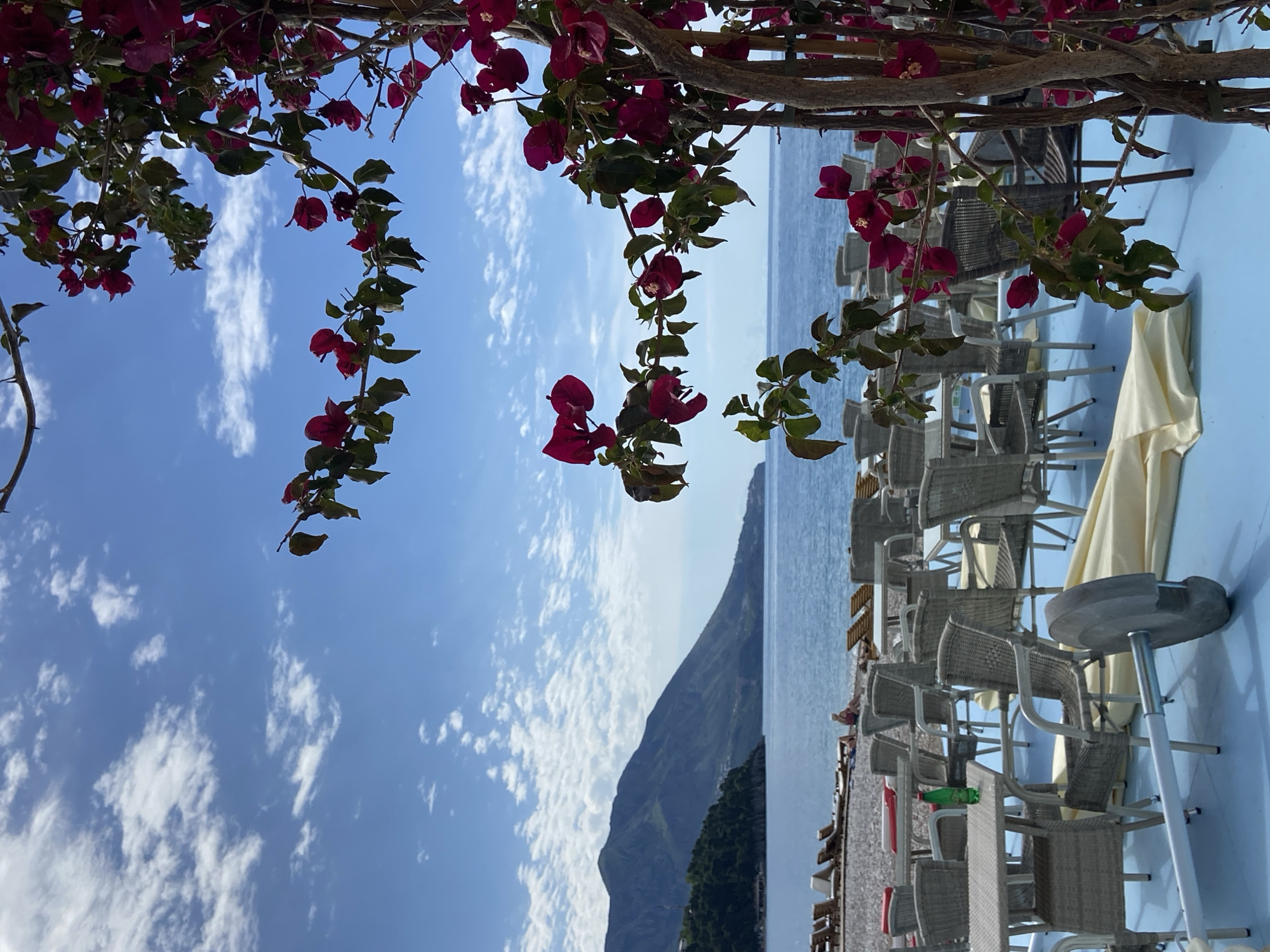 The image, rotated counter-clockwise, captures a scene at a seaside location, likely in or near Budva, Montenegro, based on the terrain and atmosphere. The foreground features red bougainvillea flowers framing the view. In the middle ground, a partially visible outdoor seating area is discernible, furnished with light-colored wicker chairs and tables, shaded by a cream-colored sun umbrella. Beyond the seating area, the Adriatic Sea stretches out to the horizon, with a mountainous landform visible in the distance. The sky is predominantly blue, interspersed with scattered white clouds. No people are clearly visible in the scene, but some people are faintly visible in the seating area and the beach.