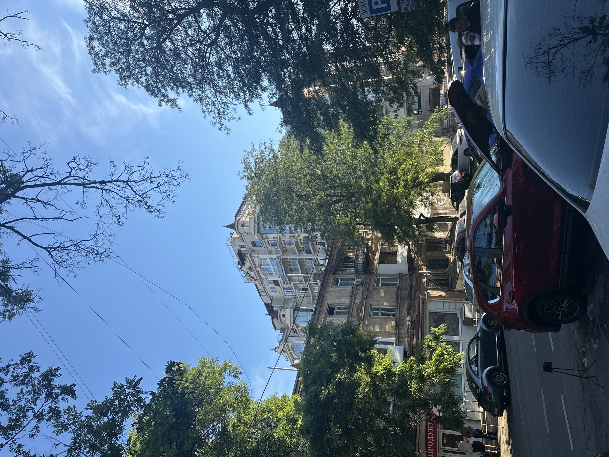 The image depicts a street scene in Odesa, Ukraine, on a sunny day, likely in the late afternoon given the long shadows. The perspective is skewed, looking upwards from near ground level. Dominating the frame are several cars parked along the side of a building. Among them is a red car in the foreground and a dark blue or black car behind it. Further down the street, more vehicles are visible.
Behind the parked cars, an old, weathered building with multiple windows and ornate architectural details stands. Adjacent to it, a taller, more modern building with a light-colored facade and numerous balconies rises into the sky. Lush green trees, some bare-branched and others in full foliage, are interspersed throughout the scene, with their branches reaching towards the bright blue sky, which is partially obscured by thin, wispy clouds.
On the sidewalk in front of a storefront, two individuals are standing, engaged in conversation. One appears to be wearing a light-colored shirt and dark pants, while the other is dressed in a darker outfit. The storefront has a red sign with Cyrillic text that partially reads "ПРОДУКЦ" which likely translates to "products". Another sign above the building entrance also displays Cyrillic text. The overall atmosphere suggests a typical urban environment in Odesa, with a mix of historical and modern architecture, verdant trees, and the presence of daily life.