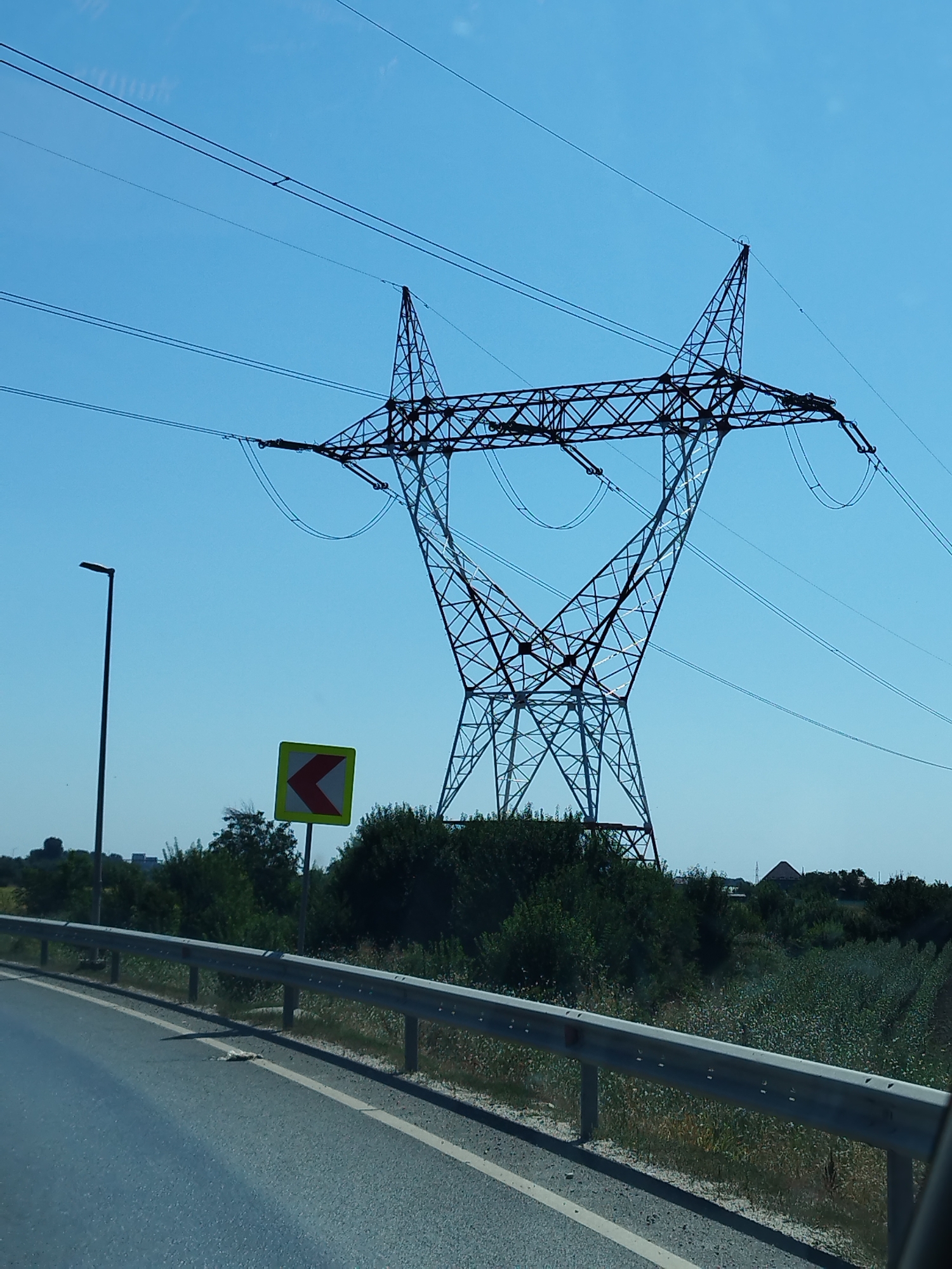 The image captures a daytime view of a transmission tower and its electrical lines against a clear blue sky. The tower is a large, lattice-style structure made of dark metal. Several electrical wires are strung between the tower and other unseen supports, stretching across the frame.

In the foreground, a road is visible, with a metal guardrail separating it from a grassy area. To the left of the road, a tall, thin lamppost stands. A traffic sign, a yellow square with a red chevron pointing left, is positioned between the lamppost and the transmission tower.

The background consists of lush green vegetation, including trees and bushes. In the far distance, a few buildings with roofs are discernible amidst the greenery, suggesting a semi-urban or rural setting. The overall scene is one of infrastructure and natural landscape under a bright sky.