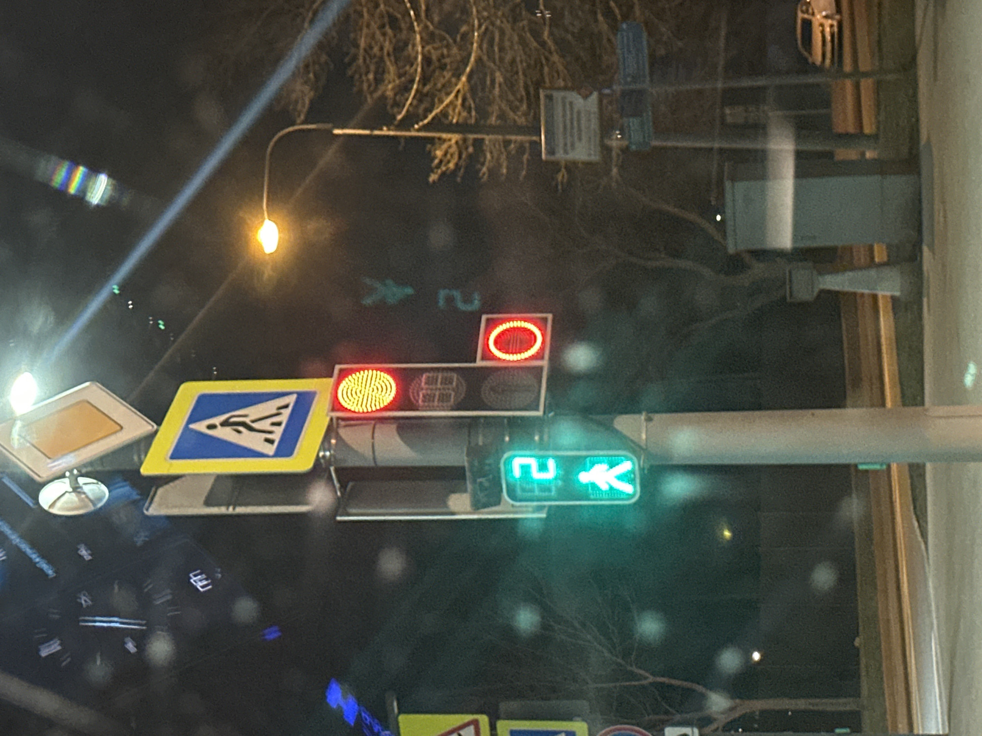 The image depicts a traffic signal in Moscow, Russia, at night. The pedestrian signal shows a green walking figure indicating it is safe to cross. Above, the traffic signal has a yellow light blinking, and a red light illuminated. Road signs indicating a pedestrian crossing are visible. Streetlights illuminate the scene. The image is likely taken from inside a vehicle due to the dashboard reflection. Bare tree branches are visible in the background.