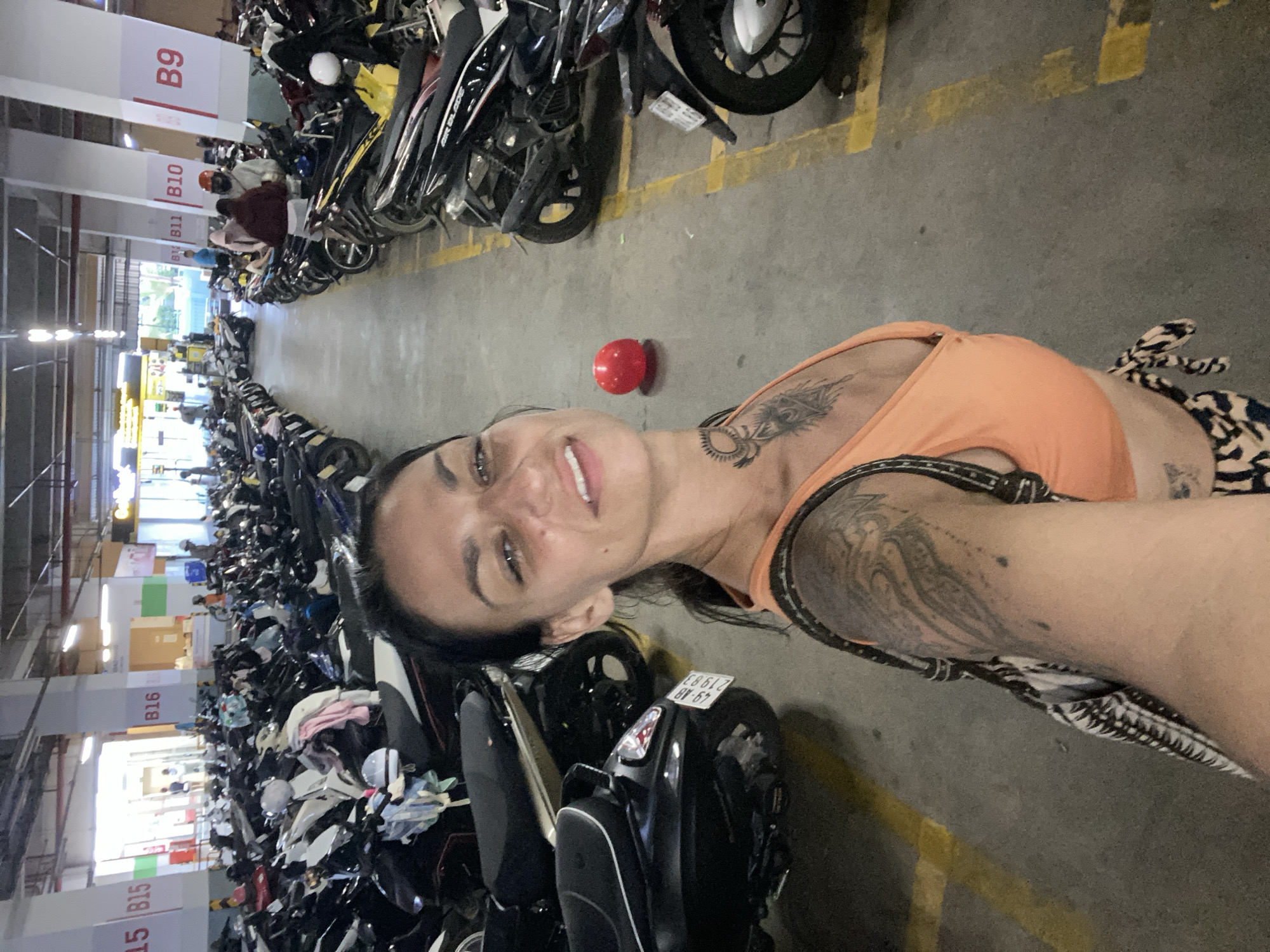 The image is a selfie taken in an indoor parking garage, likely in Phu Thuy Ward, Vietnam, given the license plate visible on a scooter that reads "49-AB 219.83", which is a Vietnamese license plate format. The foreground features a woman in an orange top, with tattoos visible on her chest and arm. She is holding a patterned bag over her shoulder. Behind her, the parking garage is filled with rows of parked motorcycles and scooters. The garage has a concrete floor with yellow painted lines and large white support columns. Overhead, fluorescent lighting illuminates the space. In the background, several people are visible, some wearing helmets and others interacting with the parked vehicles. The overall scene depicts a busy parking area for two-wheeled vehicles.