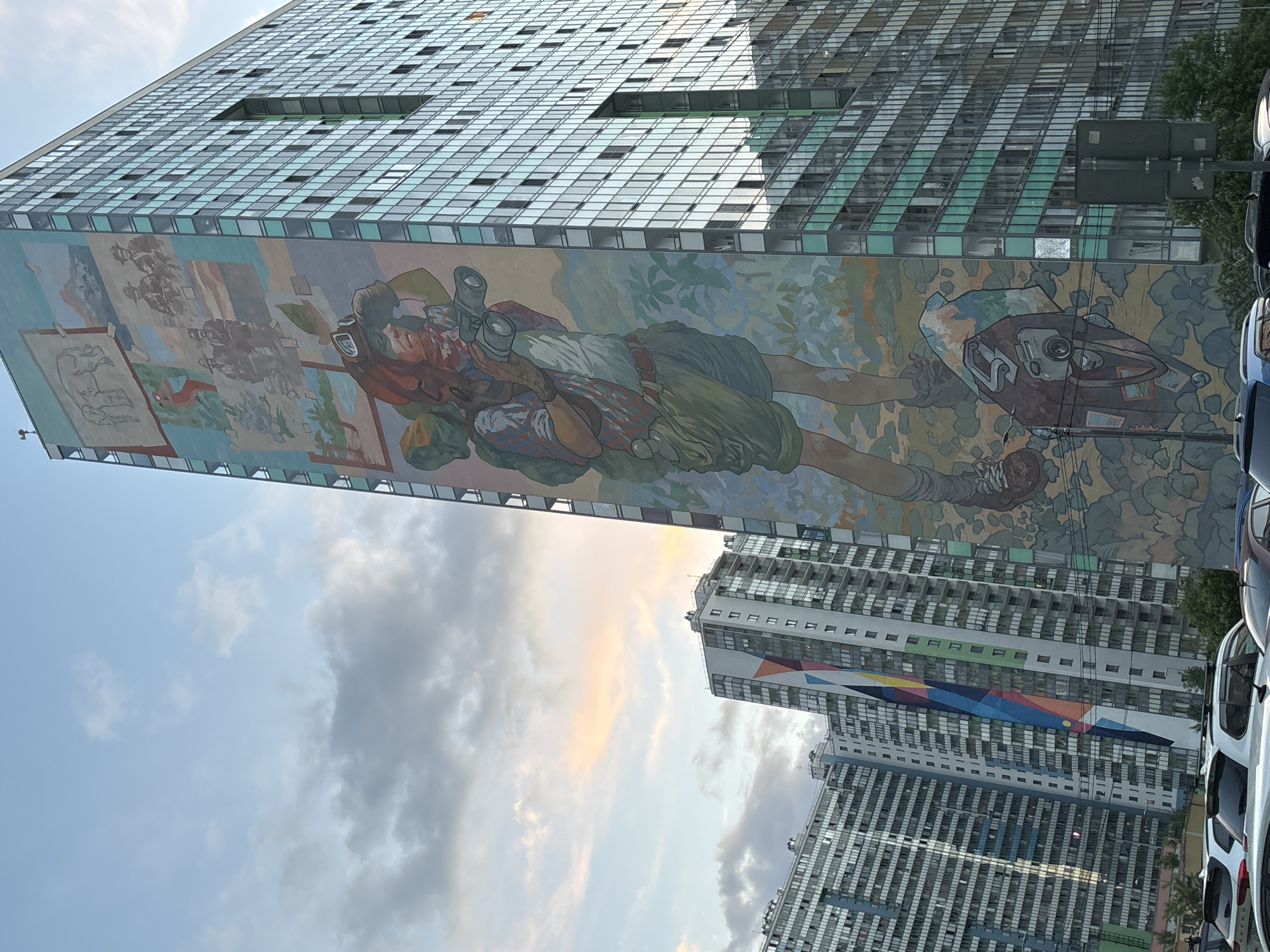 The image displays a rotated perspective of an urban scene featuring several tall buildings under a partly cloudy sky. The main focus is a large, modern building that stretches horizontally across the upper portion of the frame, indicating the image has been rotated approximately 90 degrees counter-clockwise from its natural vertical orientation. This building features a predominantly glass and metal facade, with numerous windows reflecting the sky.
A substantial mural covers a significant lower section of this main building. The mural depicts stylized human figures and abstract elements. A prominent figure is a person, possibly male, reclining or lying down, holding what appears to be binoculars or a camera lens. This figure is clad in practical attire, including a hat. To the left within the mural, there are other graphic elements, including a rough sketch of an animal (possibly an elephant or similar quadruped) and more abstract shapes, set against a background of soft, organic forms that resemble foliage or a muted landscape.
In the background, to the right and lower in the frame, are other tall residential-style buildings. One of these buildings showcases a large, vibrant geometric mural composed of intersecting triangles in various colors including blue, green, yellow, red, and black.
The sky is partly cloudy, with areas of clear blue contrasted with grey and white clouds. Hints of warm, soft light, possibly from sunrise or sunset, illuminate some of the clouds with orange and pink hues. At the very bottom right of the image, parts of several parked cars are visible, suggesting a street or parking area at ground level. Some green foliage from trees or bushes is also present near the cars and background buildings. No discernible text is visible on any of the structures or within the murals.