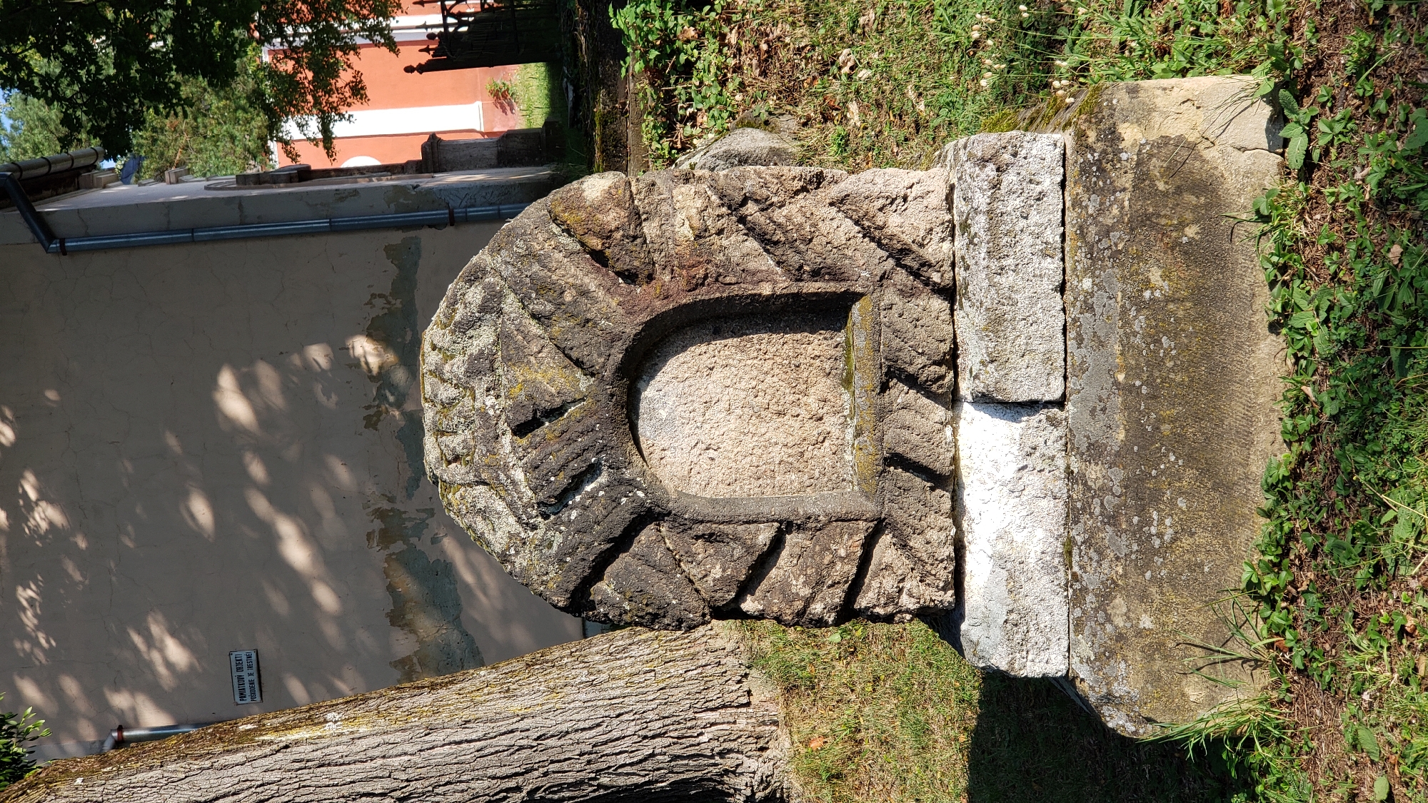 A weathered, stone monument stands in a grassy area next to a large tree and a building. The monument has a semicircular indentation in its upper section, surrounded by carved decorative patterns. The stone is rough and shows signs of age and weathering, with moss and lichen present. The monument rests on a multi-tiered stone base. In the background, a building with peeling paint and architectural details is visible, along with trees and fencing. A sign with text in Slovak is attached to the wall of the building to the left of the monument. The scene appears to be in an outdoor public space, likely a park or a historical site, based on the monument and the surrounding environment. This monument is located at the St. Nicholas Cathedral in Prešov, Slovakia.