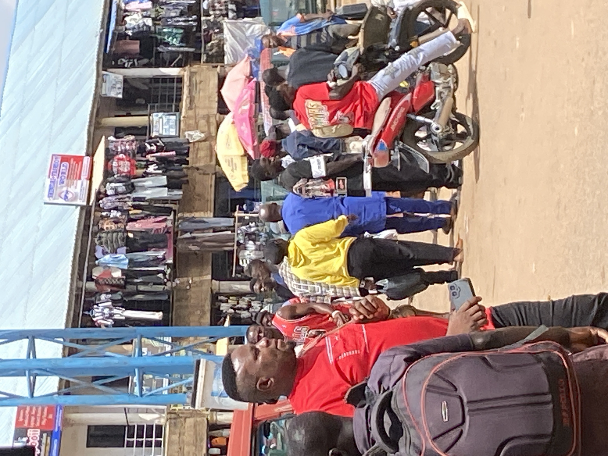This image depicts a bustling street scene in Bukuru, Nigeria, likely during daylight hours under a clear sky, suggesting fair weather. The primary subjects are a group of people, some of whom are gathered around a motorcycle that is loaded with goods. Several individuals appear to be passengers, seated or standing on the motorcycle, while others are on foot, interacting with the motorcycle or the surrounding environment.
The scene is set against a backdrop of market stalls or shops, with textiles and various merchandise displayed. The stalls are densely packed, indicating a vibrant commercial area. One stall has a prominent red sign with white text that reads "QUANTUM WORLD".
The people are dressed in casual attire. One man in the foreground is wearing a red t-shirt, and another is wearing a yellow jacket over a plaid shirt. The presence of backpacks and luggage suggests travel or transport of goods. The overall atmosphere appears to be one of movement and activity, characteristic of a busy marketplace. The people's expressions are largely neutral, though the man in the foreground seems to be looking up and to his left. The image captures a moment in the daily life of this Nigerian community, highlighting trade and transportation.