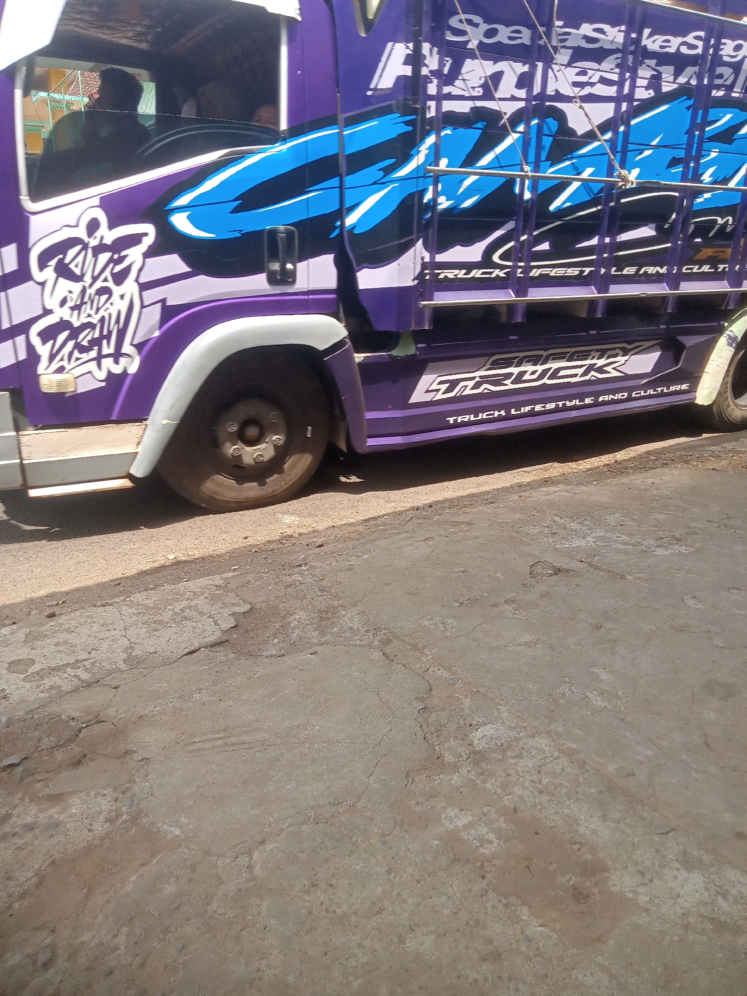 This is a medium shot, eye-level photograph of the side of a purple and white truck parked on a dirt and concrete surface, likely in a street in Pule, Indonesia. The truck is decorated with graphic designs and text. On the side of the truck, there are bold, stylized graphics in blue, white, and black. The text "RIDE AND DASH" is prominently displayed in a graffiti-like font. Further back, the text "TRUCK LIFESTYLE AND CULTURE" is visible. The truck has a white fender over its rear wheel, which is visible. The interior of the truck cab is partially visible, showing at least two people inside, one in the driver's seat. The ground in front of the truck is a mix of rough concrete with cracks and patches of dirt and gravel. The lighting suggests it is daytime.