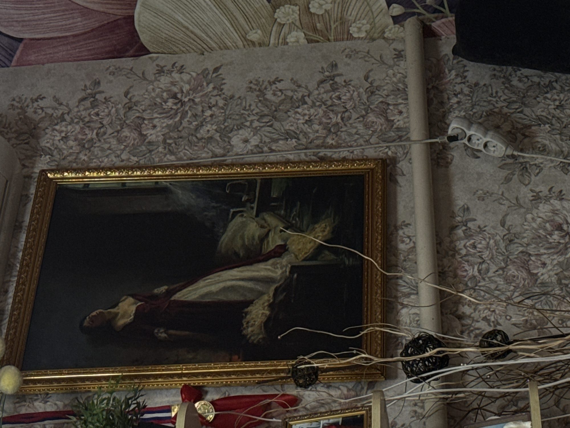 A framed painting hangs on a wall covered in floral wallpaper. The painting depicts a reclining woman. A white extension cord is plugged into the wall near a vertical pipe. There are decorative branches with round, woven ornaments in front of the painting. Below the painting are more decorations and the top of another framed object. The location is Unknown, Unknown.