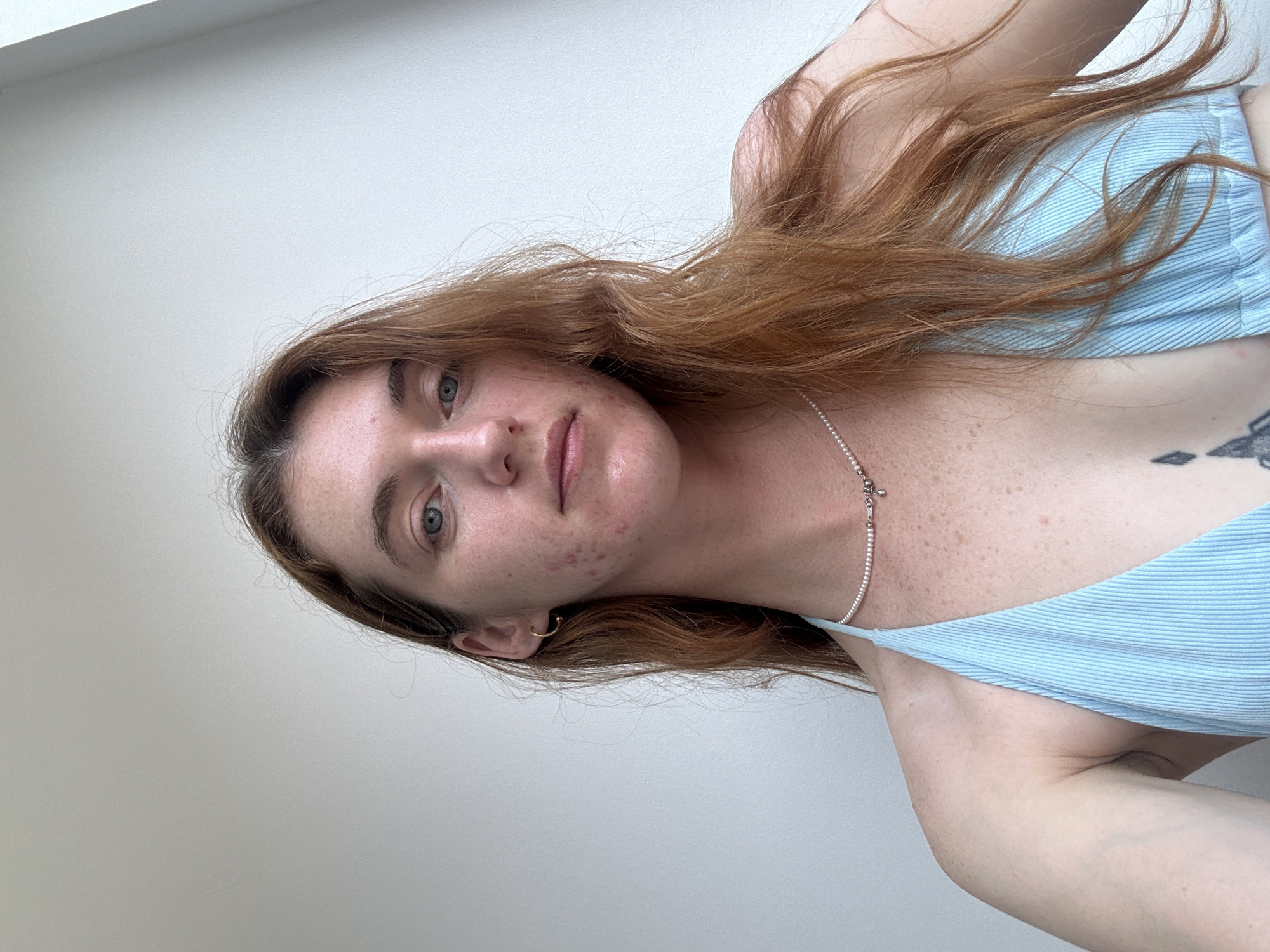 A medium shot depicts a fair-skinned woman with long, red-brown hair. She is wearing a light blue striped top. A silver necklace adorns her neck. Her skin displays some freckles and minor acne scarring. The background is a plain, light-colored wall. The image appears to have been taken indoors. There is no indication of the location being Baan Chaweng, Thailand.