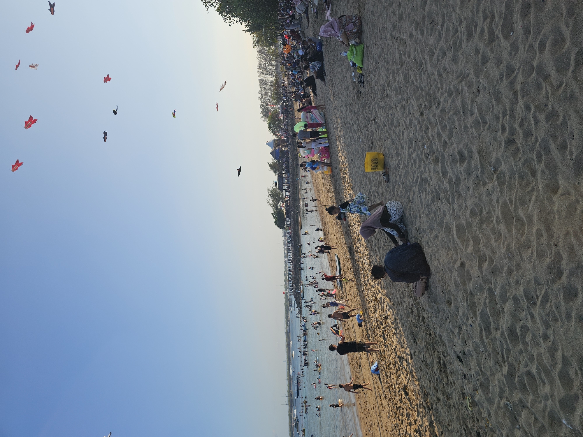 This is a wide-angle, eye-level shot of a crowded beach in Denpasar, Indonesia. The sand is a light brown, with some ripples and footprints visible. In the background, the ocean is calm and a light blue, with many boats and people swimming. There are also structures in the distance, including buildings and what appears to be a pier. The sky is a clear, light blue with a few white clouds.
Many people are on the beach and in the water, engaged in various activities such as swimming, playing, and relaxing. Some are sitting on the sand, while others are walking or standing. There are also several kites flying in the sky, adding to the festive atmosphere.
The people on the beach are diverse in age and gender. Many are wearing casual beach attire. Some are dressed in traditional Indonesian clothing, such as headscarves. The people appear to be enjoying their time at the beach.
The overall impression of the image is one of a vibrant and lively beach scene, likely a popular tourist destination.