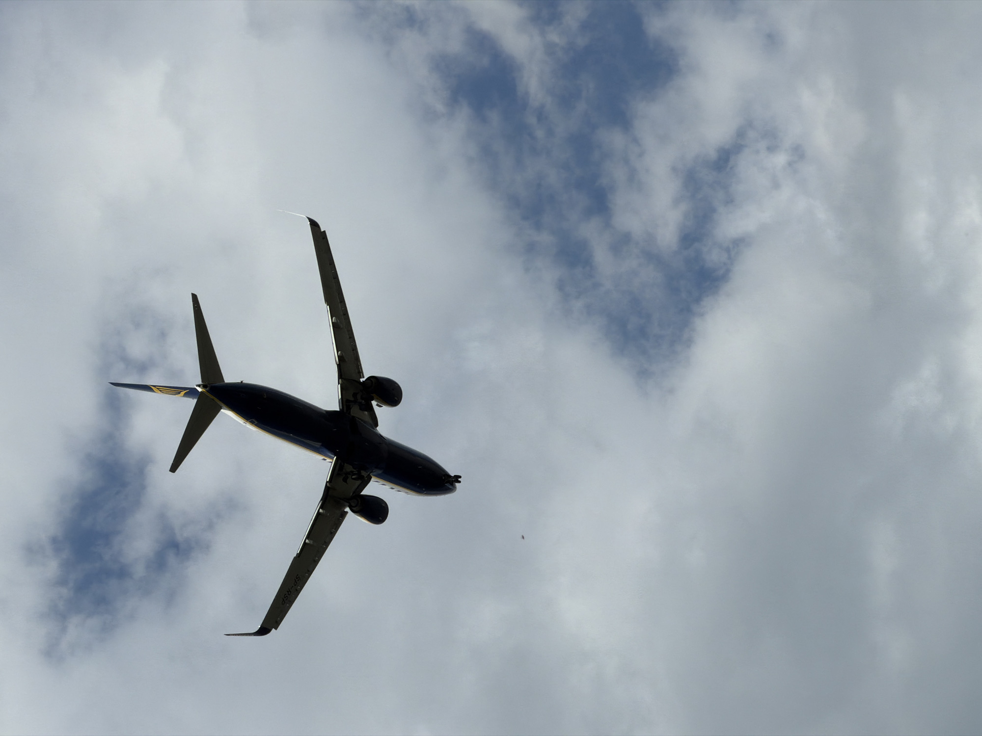 An airplane, identified by the logo on its tail as likely Ryanair, is captured from below, ascending into a sky filled with clouds. The aircraft is predominantly dark blue with yellow accents, and its landing gear is extended. The registration number "SP-RSP" is visible on the wing. The scene is set against a backdrop of a cloudy sky, with patches of blue sky visible through the clouds. The photo was taken near Burgas, Bulgaria.