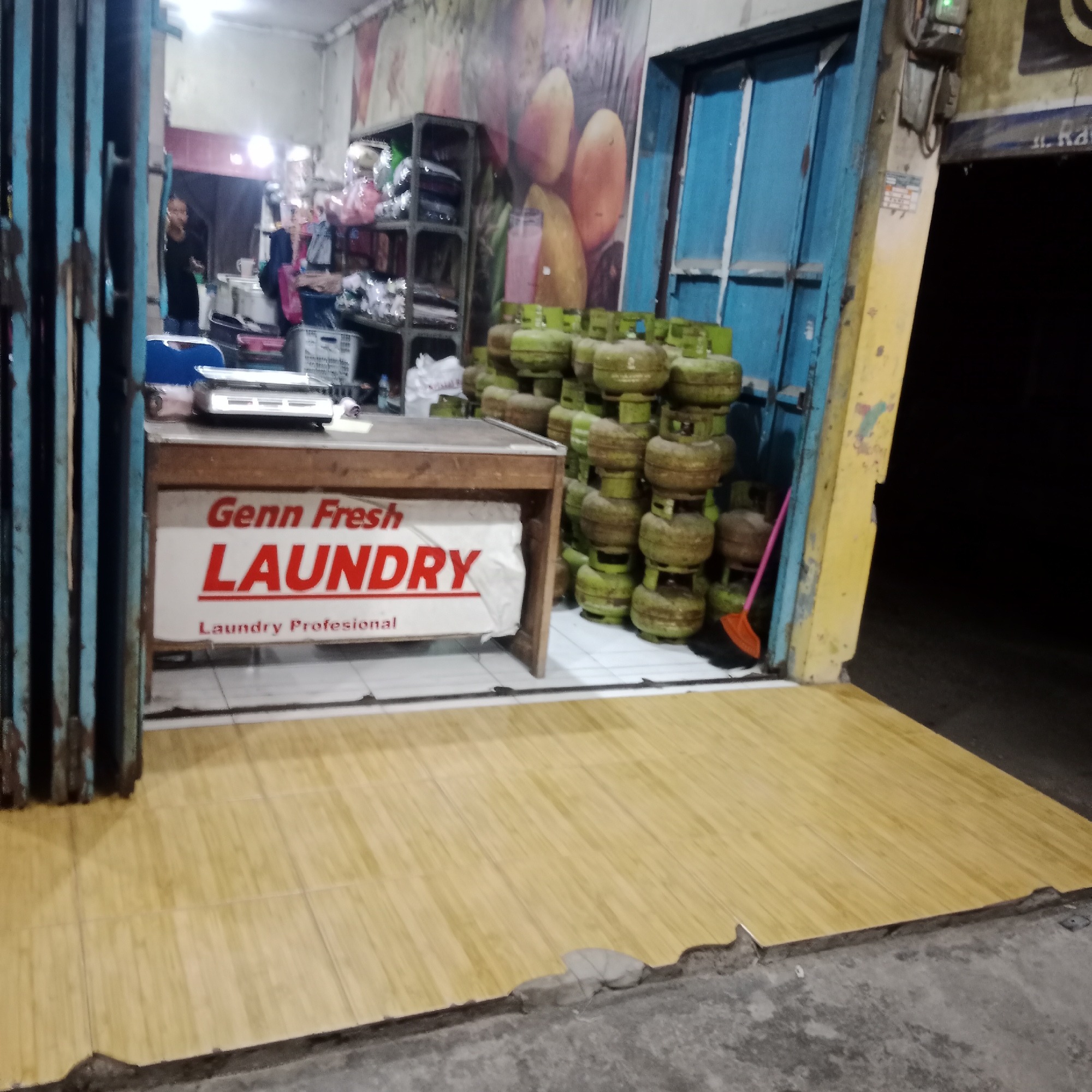 The image depicts the exterior of a small business, possibly a laundry service, located in Tegowanu Kulon, Indonesia. In the foreground, a wooden counter with a sign reading "Genn Fresh LAUNDRY" and "Laundry Profesional" is visible. Behind the counter, a stack of green propane tanks is arranged. To the left, a person is partially visible behind a blue gate. The shop's facade features a large mural of fruit and a blue door. The ground is tiled with light brown square tiles. The scene appears to be at dusk or night, with artificial lighting illuminating the area.
