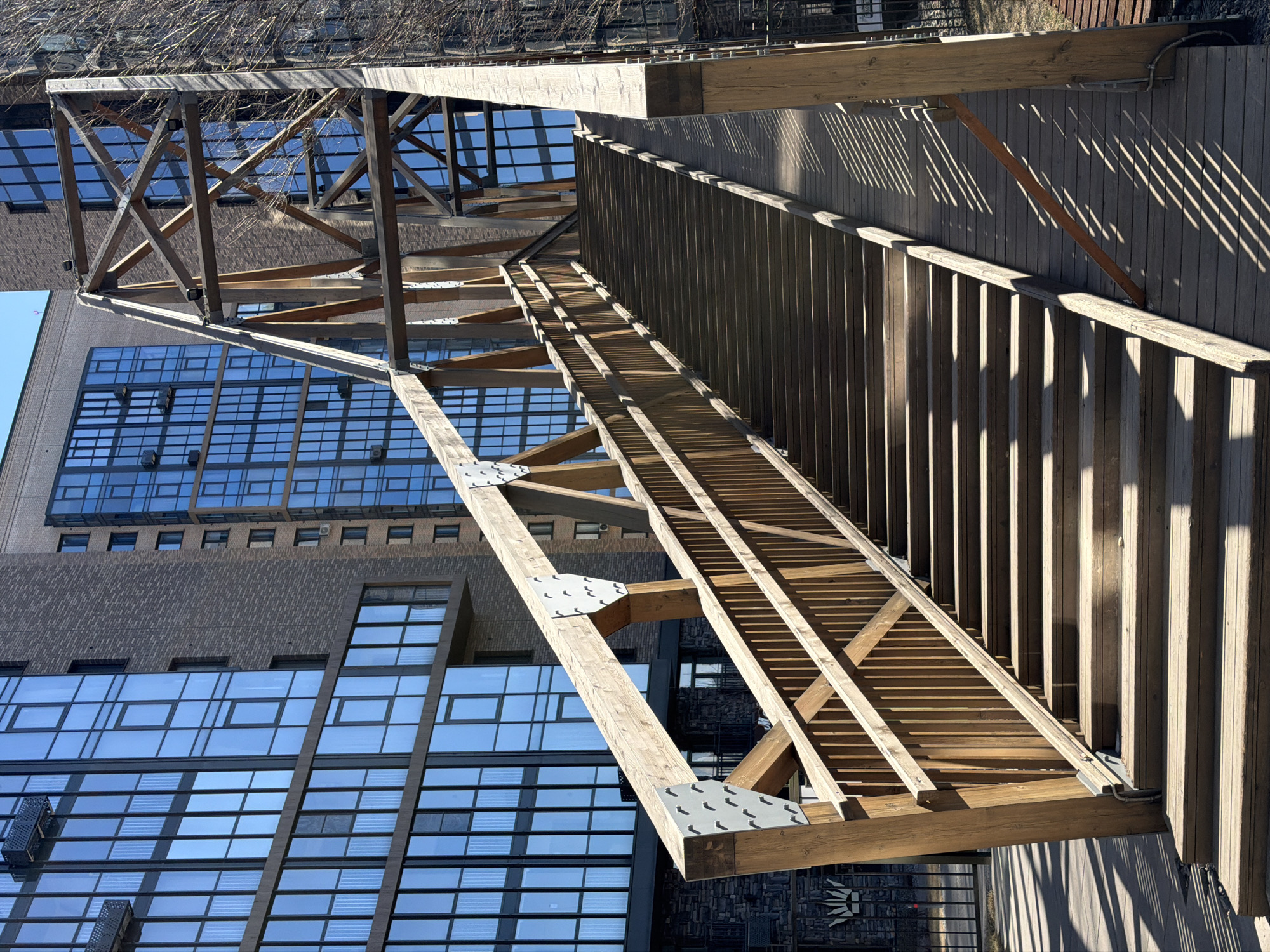 The image shows a wooden pedestrian overpass in an urban environment. The overpass is constructed with exposed wooden beams and metal connectors. A walkway with a slatted wooden fence runs along one side. In the background, there are modern buildings with glass curtain walls reflecting the sky. Based on the architectural style and urban setting, the location is likely a contemporary development within a city. The overpass structure casts strong shadows due to the angle of the sunlight. There are no people visible in the frame. Without specific knowledge of Krasnoyarsk's architecture or notable landmarks in the image, the exact location within the city cannot be determined.