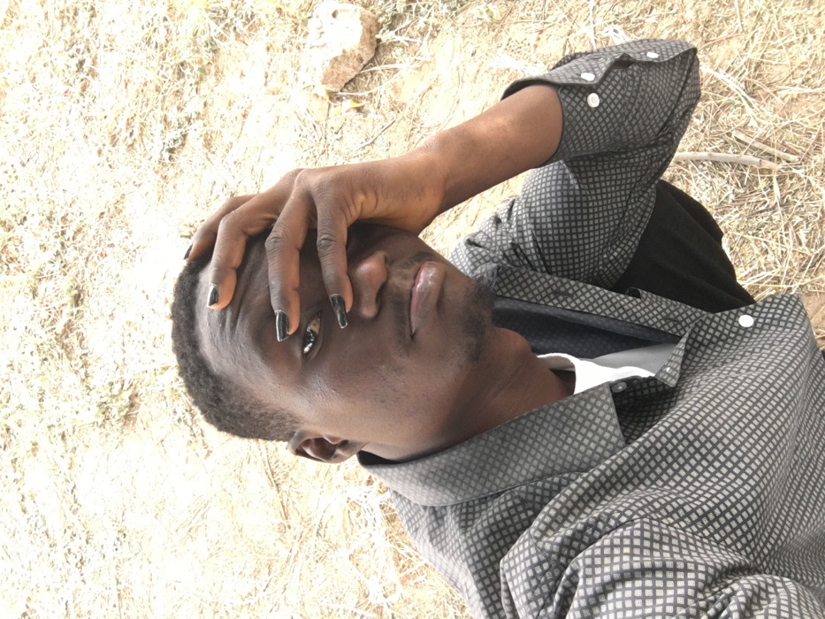 A dark-skinned person lies on dry, sunlit ground. The individual wears a patterned gray shirt over a white undershirt. Their black-painted fingernails are visible as a hand rests on their forehead. A slight beard and mustache are present. The person looks up, slightly sideways to the camera. The background consists of dry earth, scattered leaves, and a small rock.